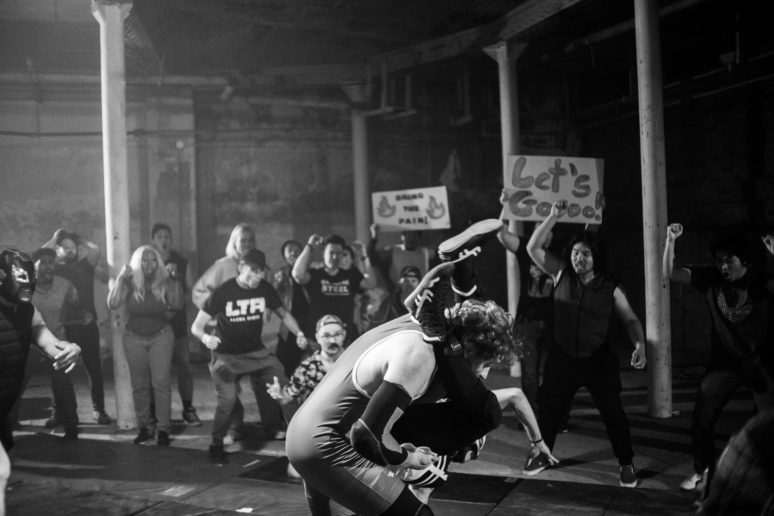Black and white photo of a wrestling match with a cheering crowd holding signs.