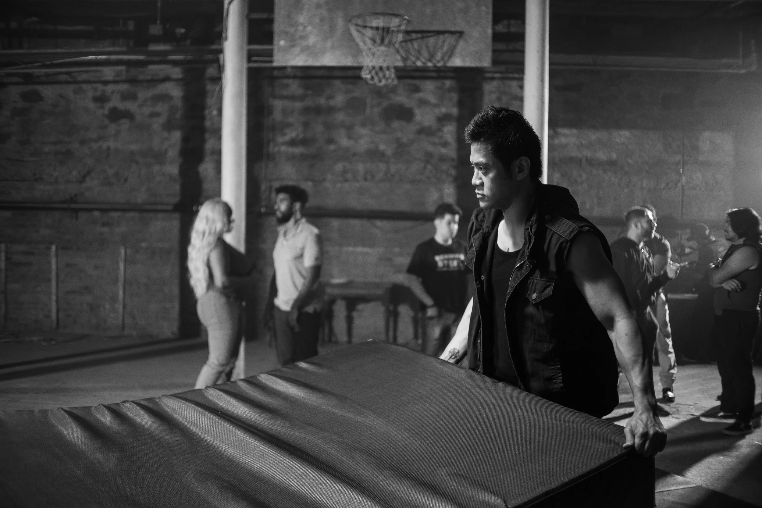 Black and white photo of a group of people in a dimly lit gymnasium. In the foreground, a man wearing a sleeveless jacket is moving a large pad. Other individuals are conversing in the background, and a basketball net is visible.
