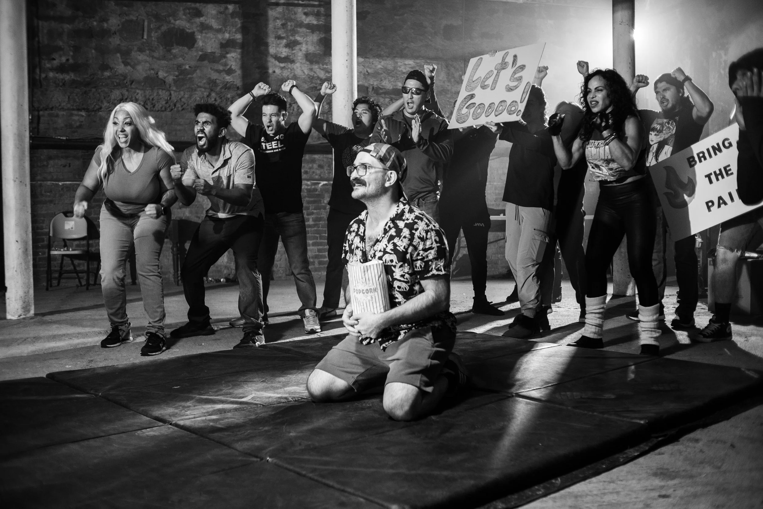 Black and white image of a group of people cheering around a man kneeling on a mat with a popcorn container. The crowd holds signs like 'Let's Go' and 'Bring the Pain,' appearing enthusiastic and supportive.