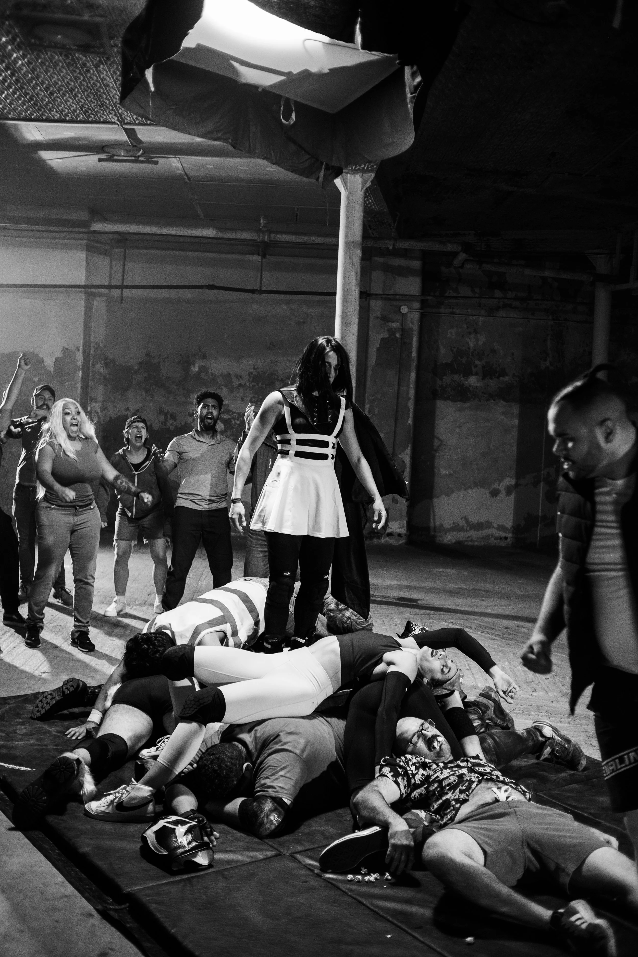 Black and white scene showing a person in a theatrical outfit standing triumphantly on a pile of people lying on the ground, surrounded by cheering individuals in an industrial setting.