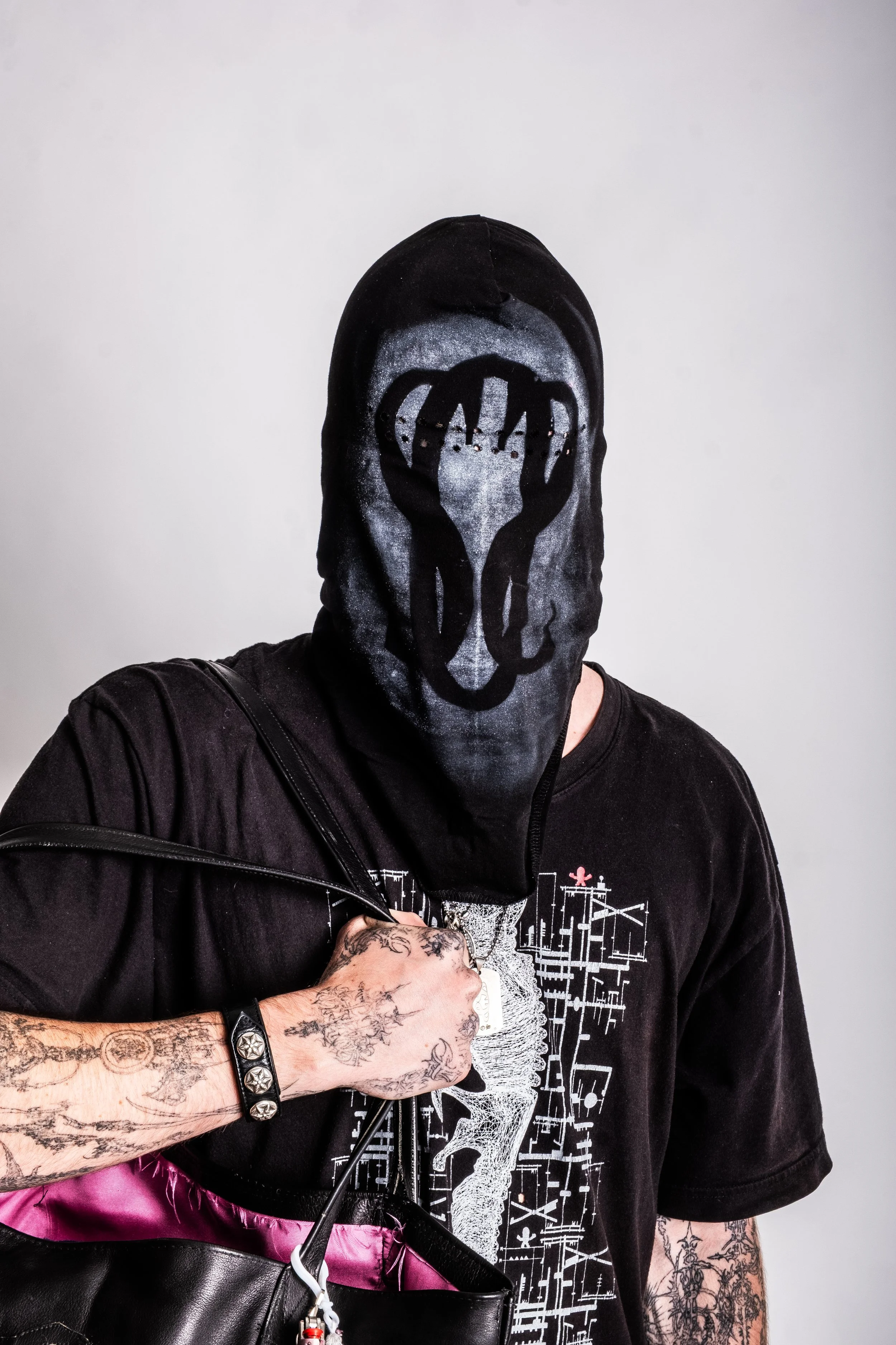 Person wearing a black mask with a printed design, holding a black handbag, and wearing a black T-shirt and bracelet with tattoos visible on the arm.