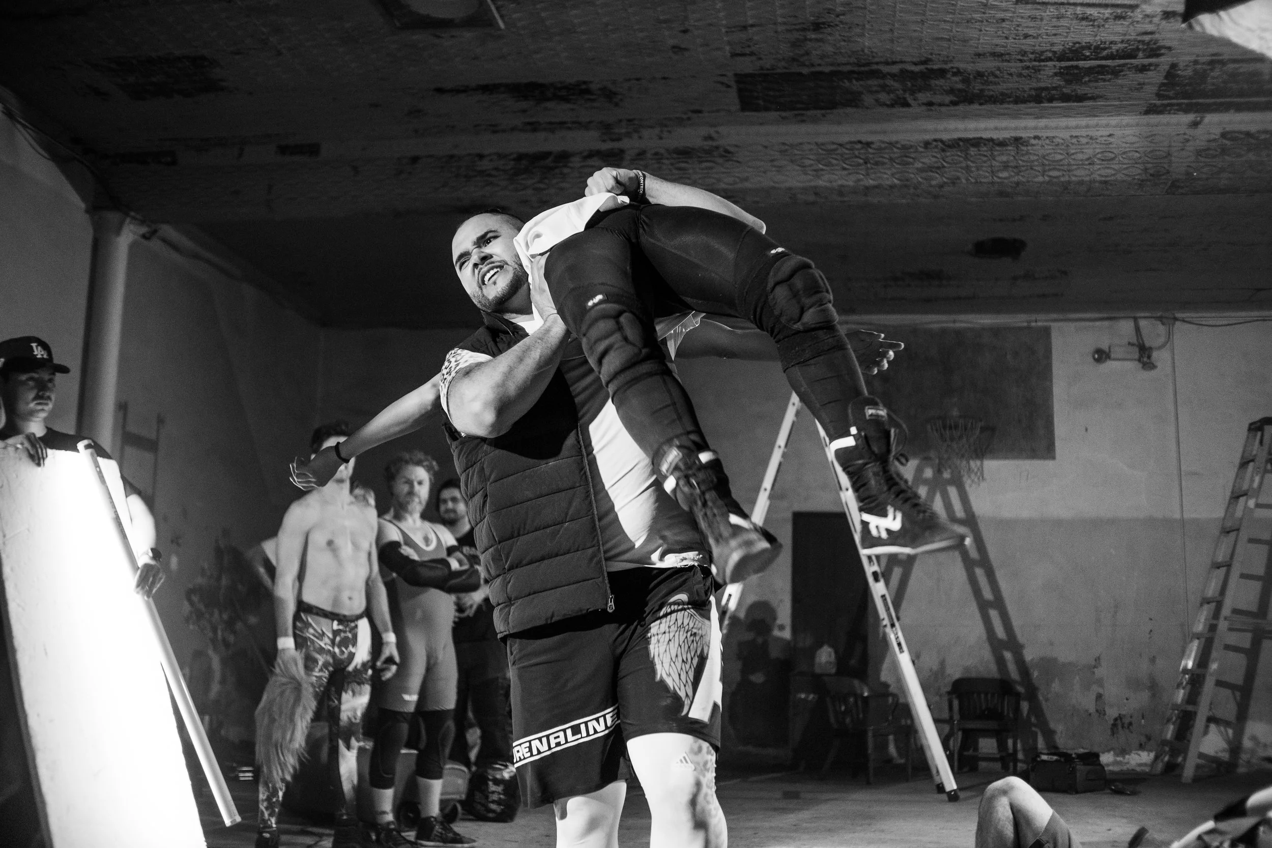 Black and white image of a wrestling event in a gym, featuring a man lifting another wrestler over his shoulder amidst onlookers, with ladders and chairs in the background.