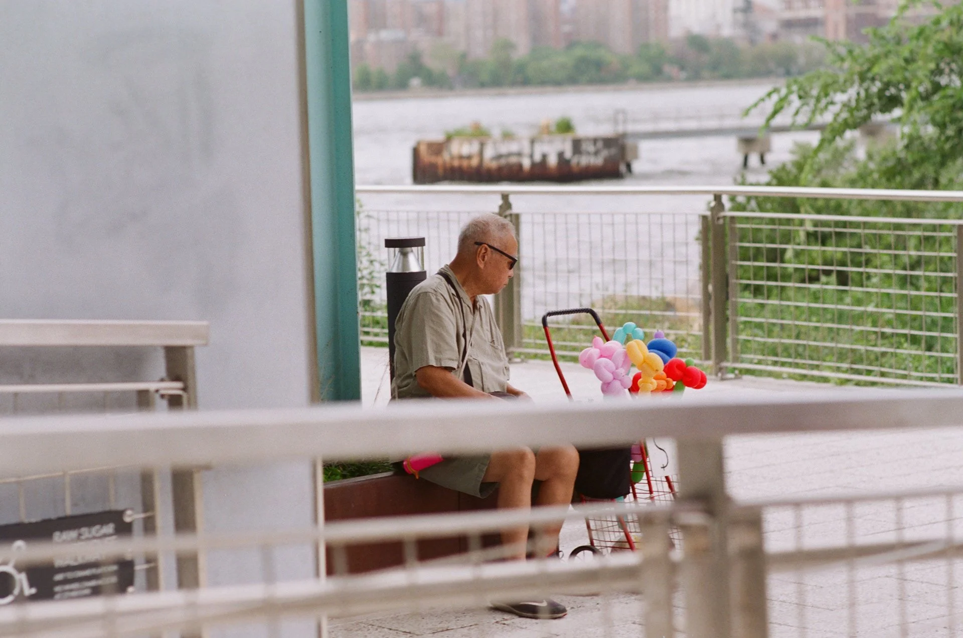 Man sitting on a bench with colorful balloons near a riverfront fence.