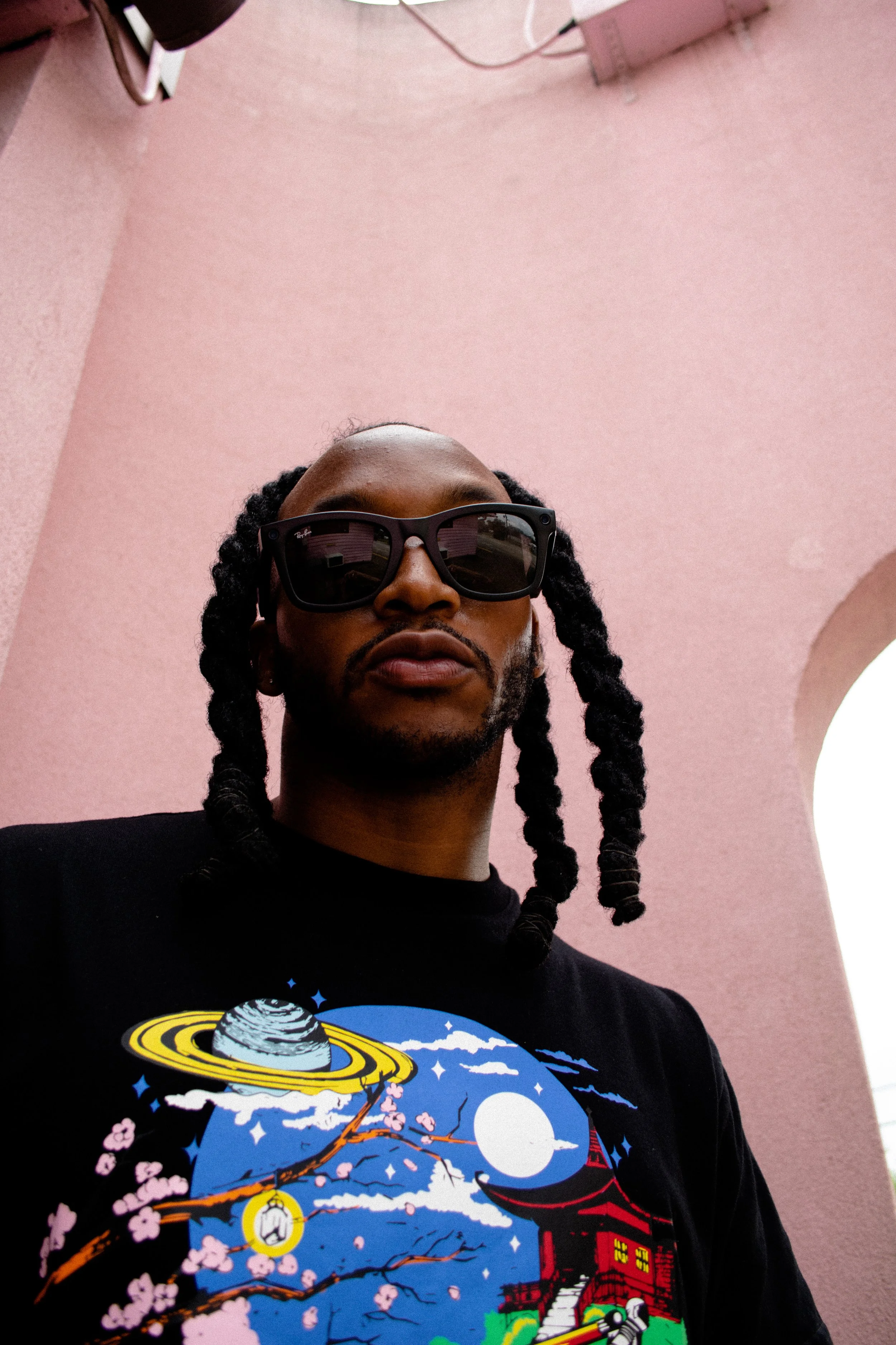 Person wearing sunglasses and a black shirt with colorful design, against a pink background.