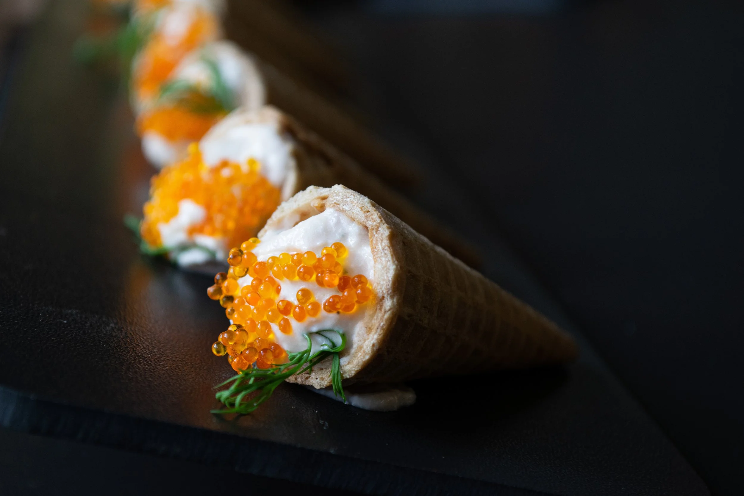 Waffle cone with cream cheese, orange caviar, and dill garnish on dark surface.
