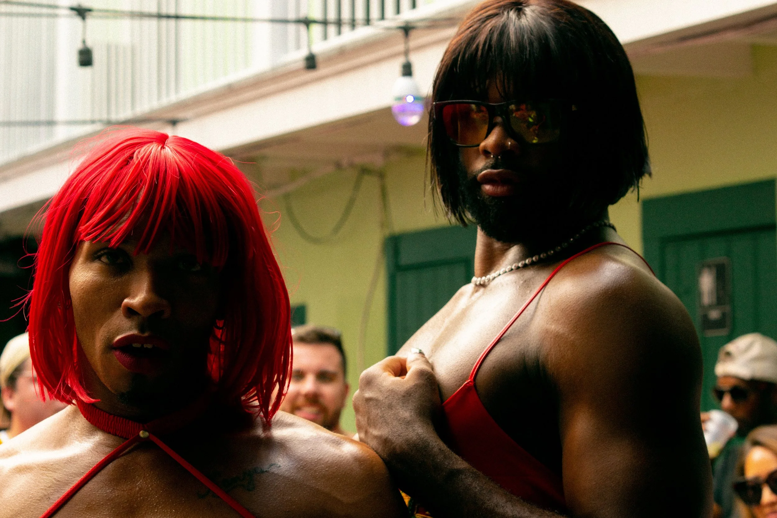 Two individuals wearing red costumes and wigs stand in an outdoor setting. One has a bright red wig, and the other has a dark wig and sunglasses. Both display strong, confident poses.