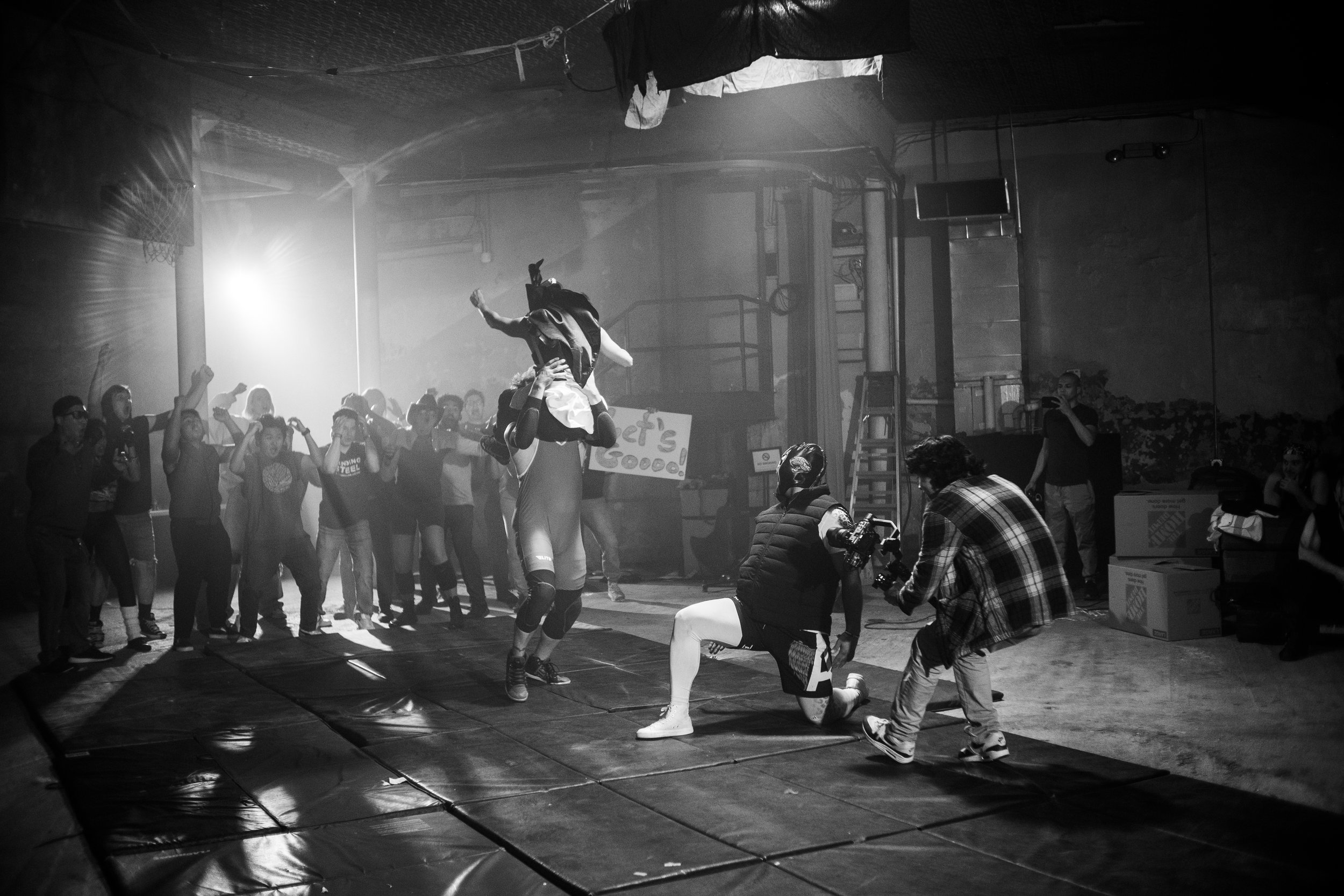 Black and white photo of a wrestling match in an underground venue, with a crowd cheering. Wrestlers in masks perform a move in the center. A cameraman kneels to capture the action. Dim lighting and industrial surroundings enhance the intense atmosph