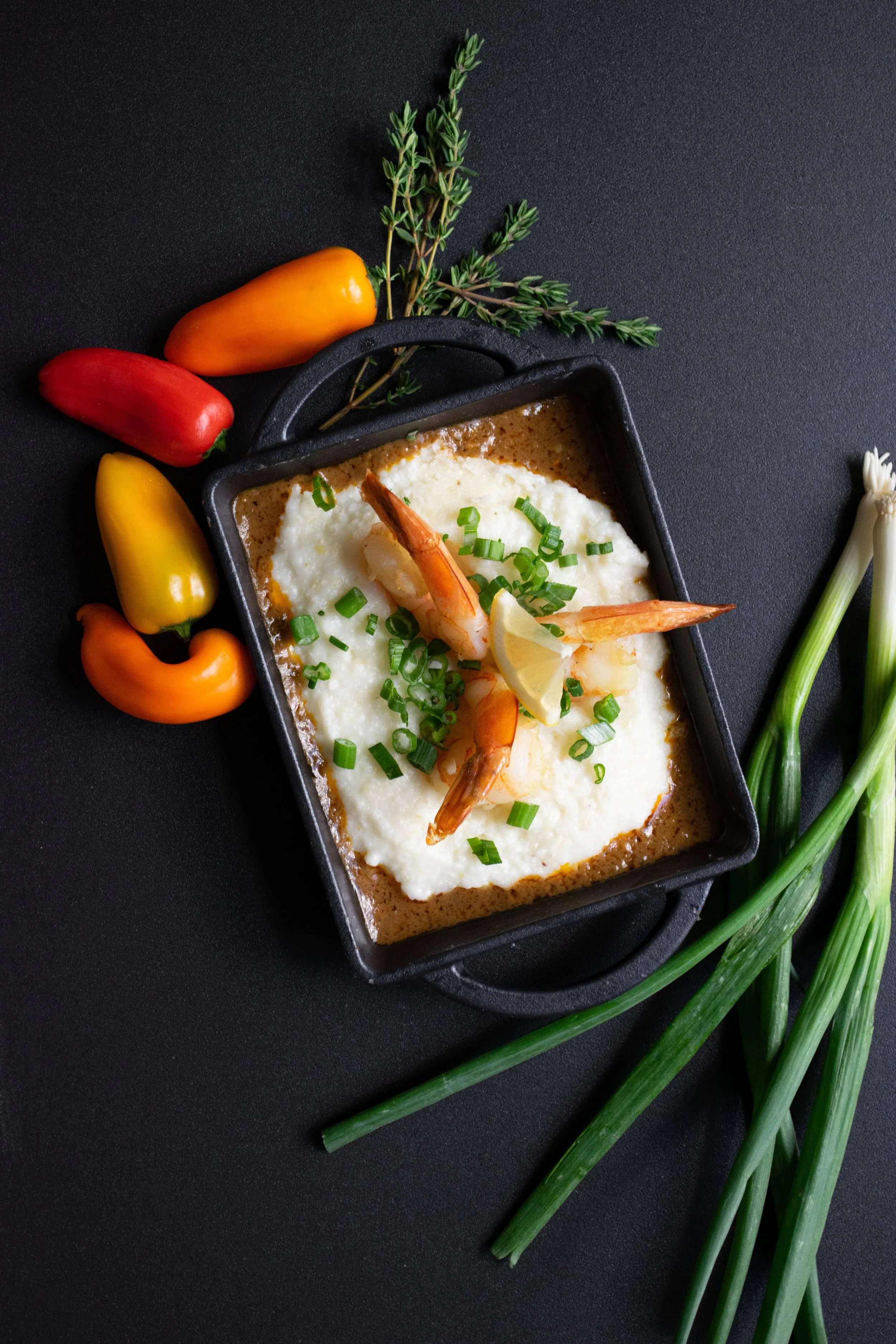 Shrimp and grits in a black dish with sliced green onions, lemon wedge, and thyme, surrounded by colorful bell peppers and green onions on a dark surface.