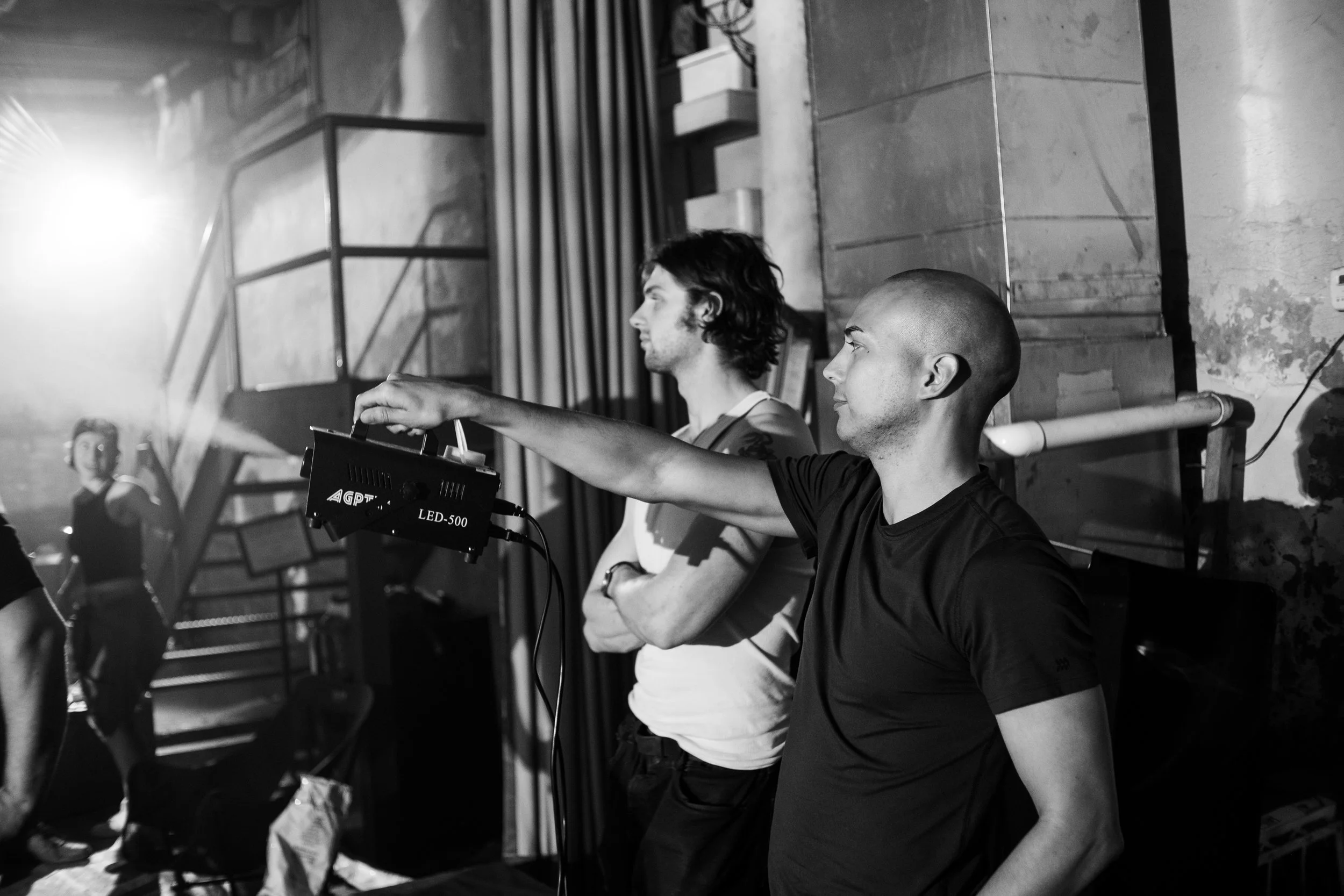 Men working behind the scenes of a stage production, adjusting a spotlight in a dimly lit environment.