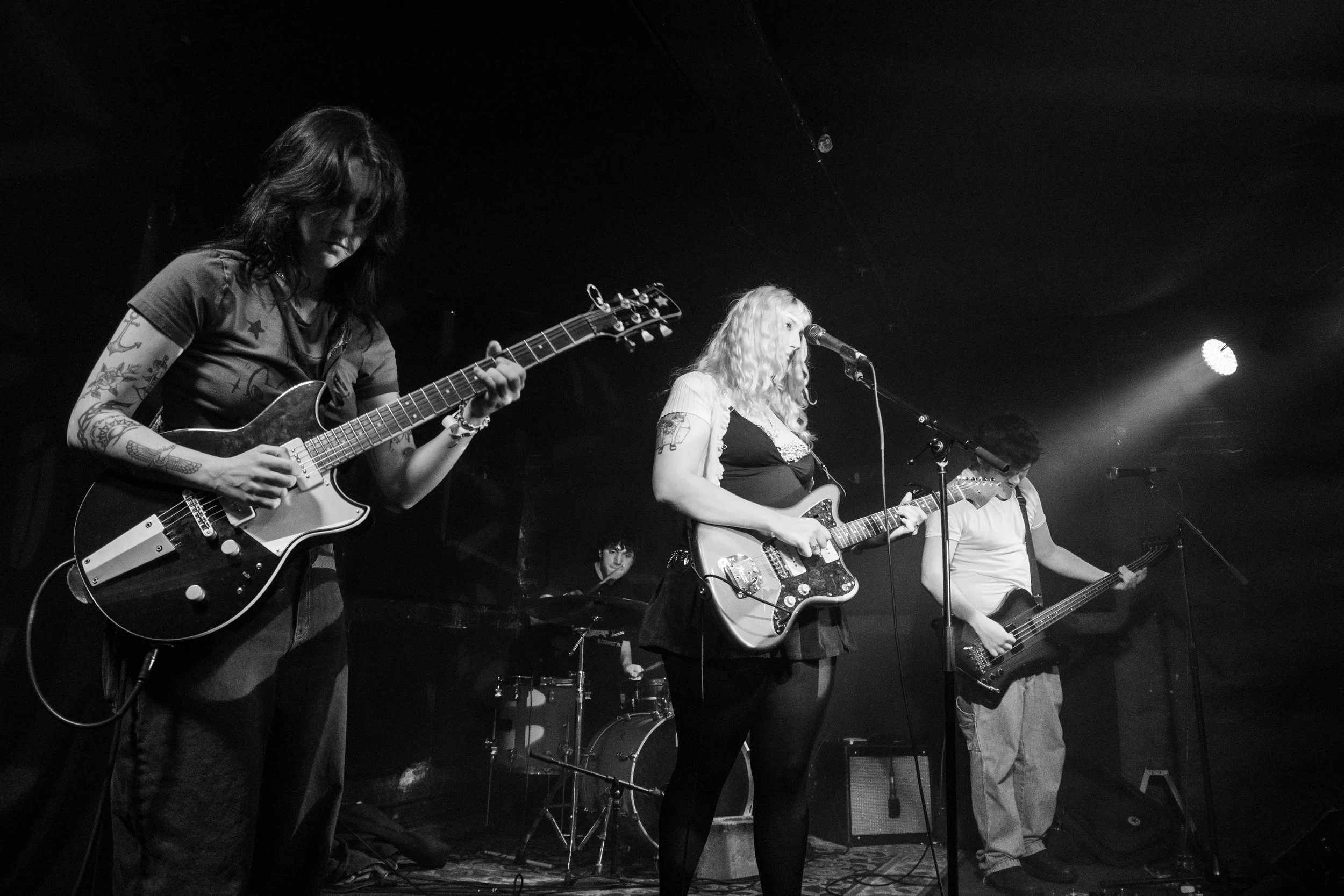 Black and white photo of a rock band performing on stage with two guitarists and a drummer.