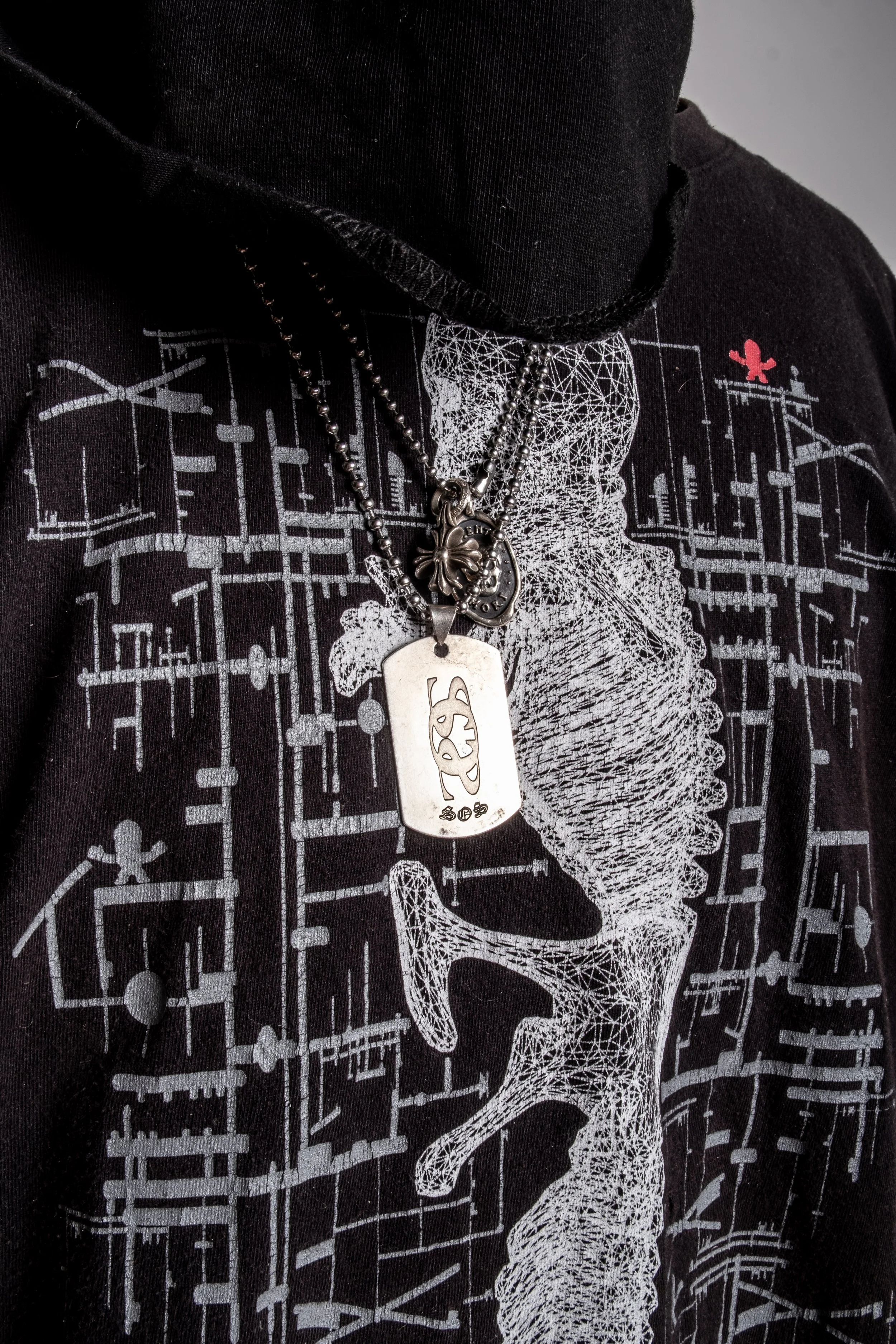Close-up of a black hoodie featuring complex white geometric and skeletal patterns, adorned with layered metal necklaces, one with a rectangular pendant inscribed with symbols.