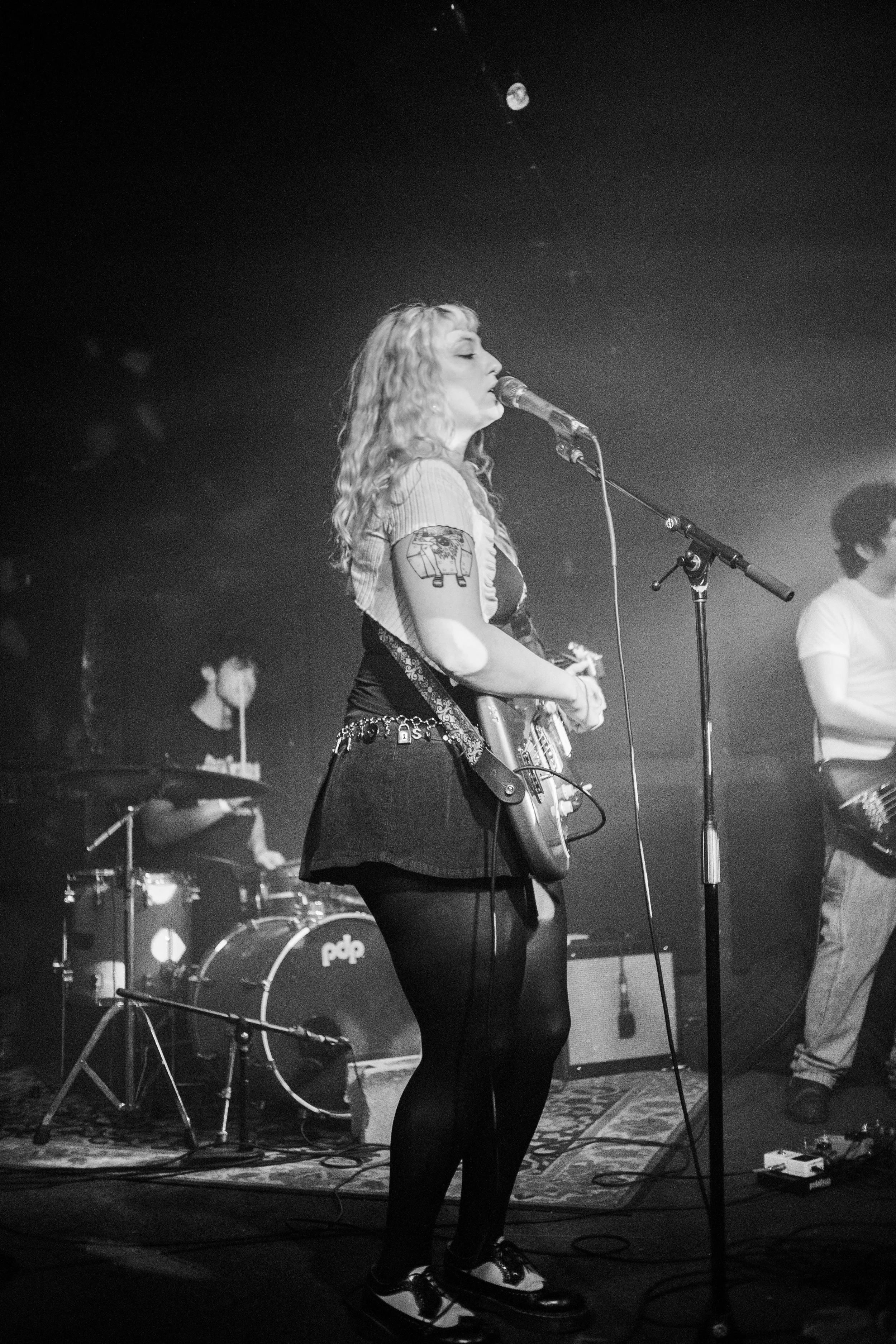 Black and white photo of a female guitarist and singer performing on stage with a drummer in the background. The guitarist is playing an electric guitar and singing into a microphone.