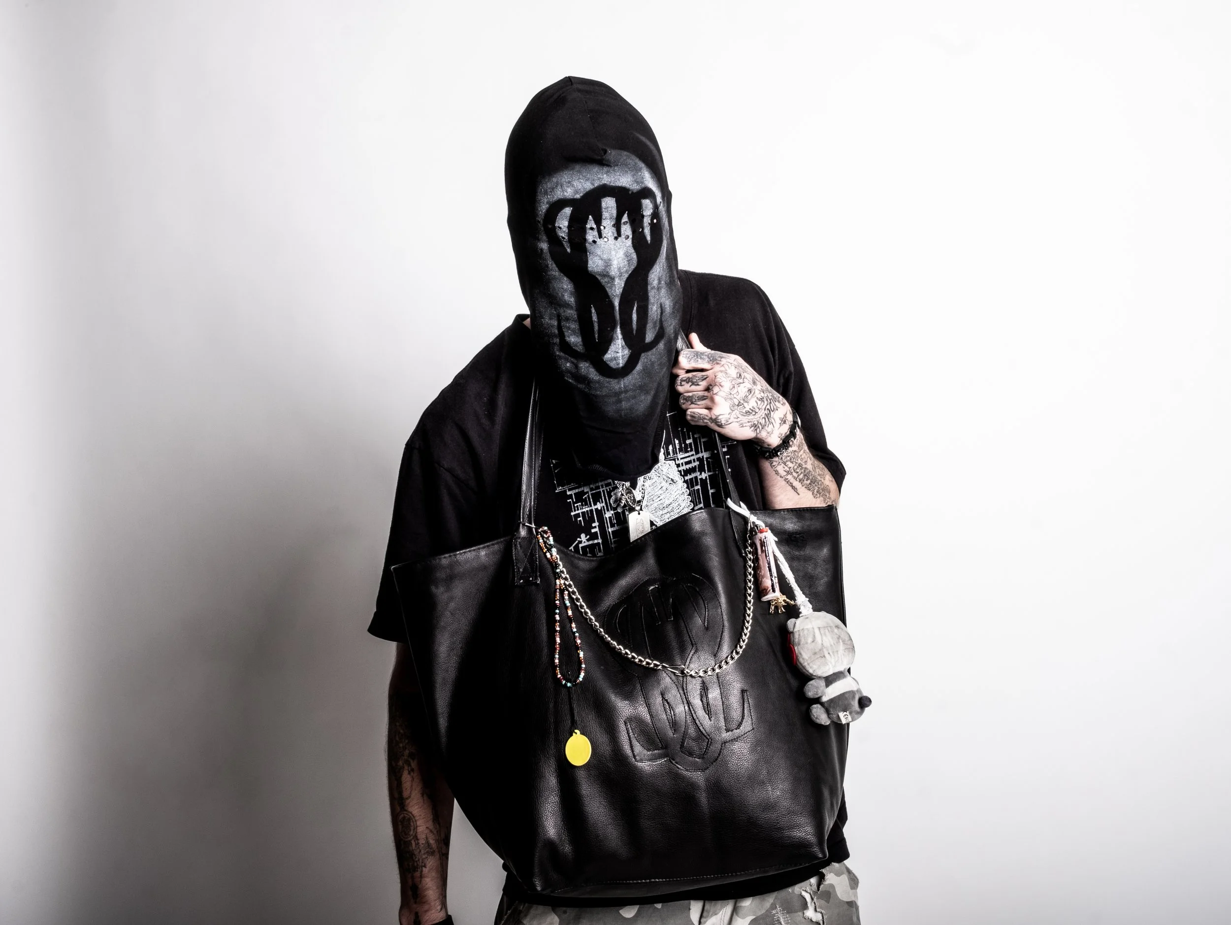 Person wearing a black hooded mask and shirt, holding a large black leather bag with a chain and charms. Tattoos are visible on their arms.