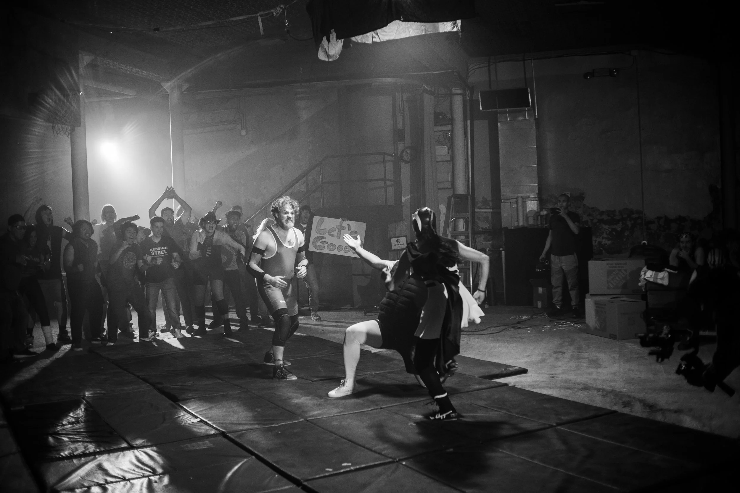 Black and white photo of two wrestlers in action in a crowded industrial setting with cheering spectators.