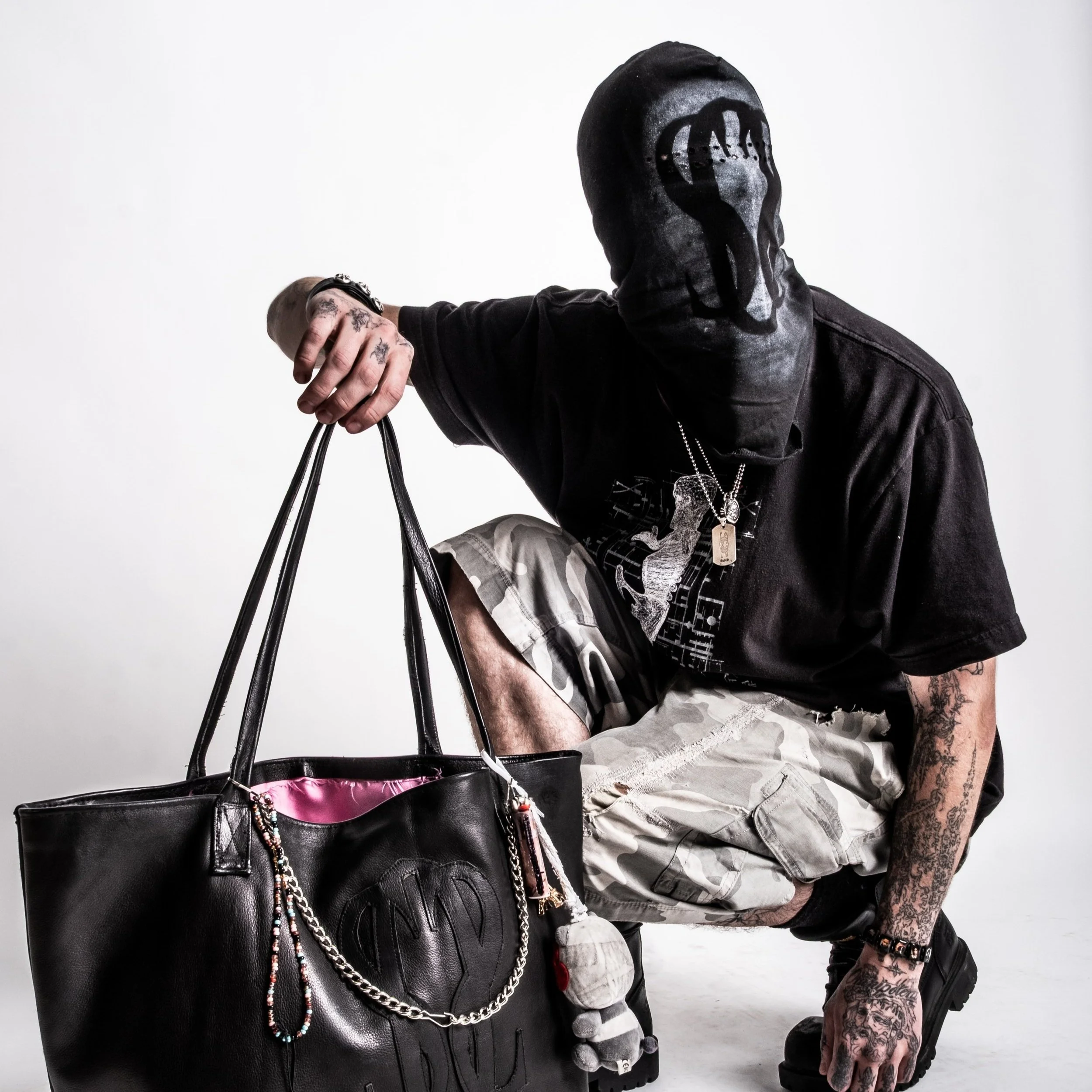 A person in a black hood and casual clothing, holding a large black tote bag with decorative chains and patterns, crouching in a white studio setting.