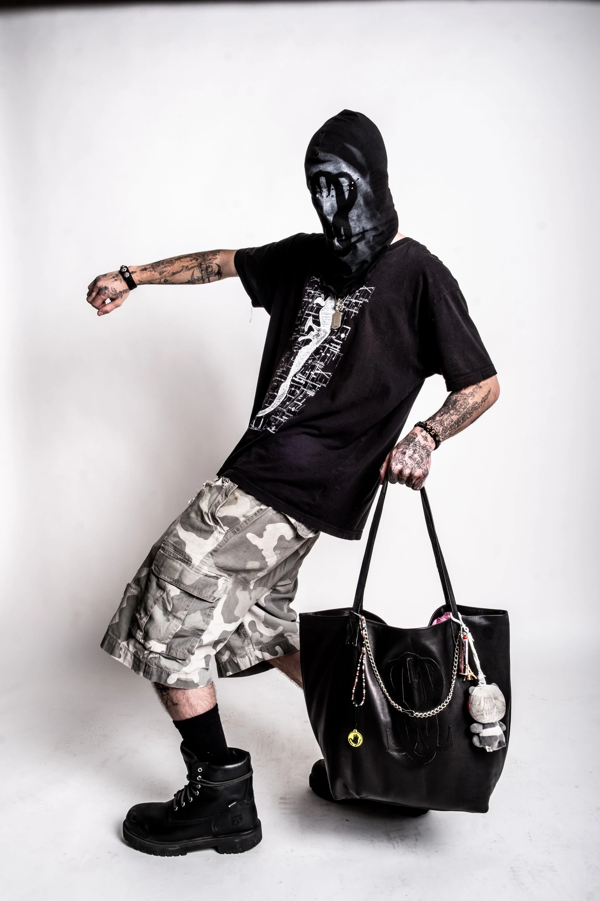 Person in camo shorts, black T-shirt, and boots, with a hood covering face, holding a large black bag in a studio setting.