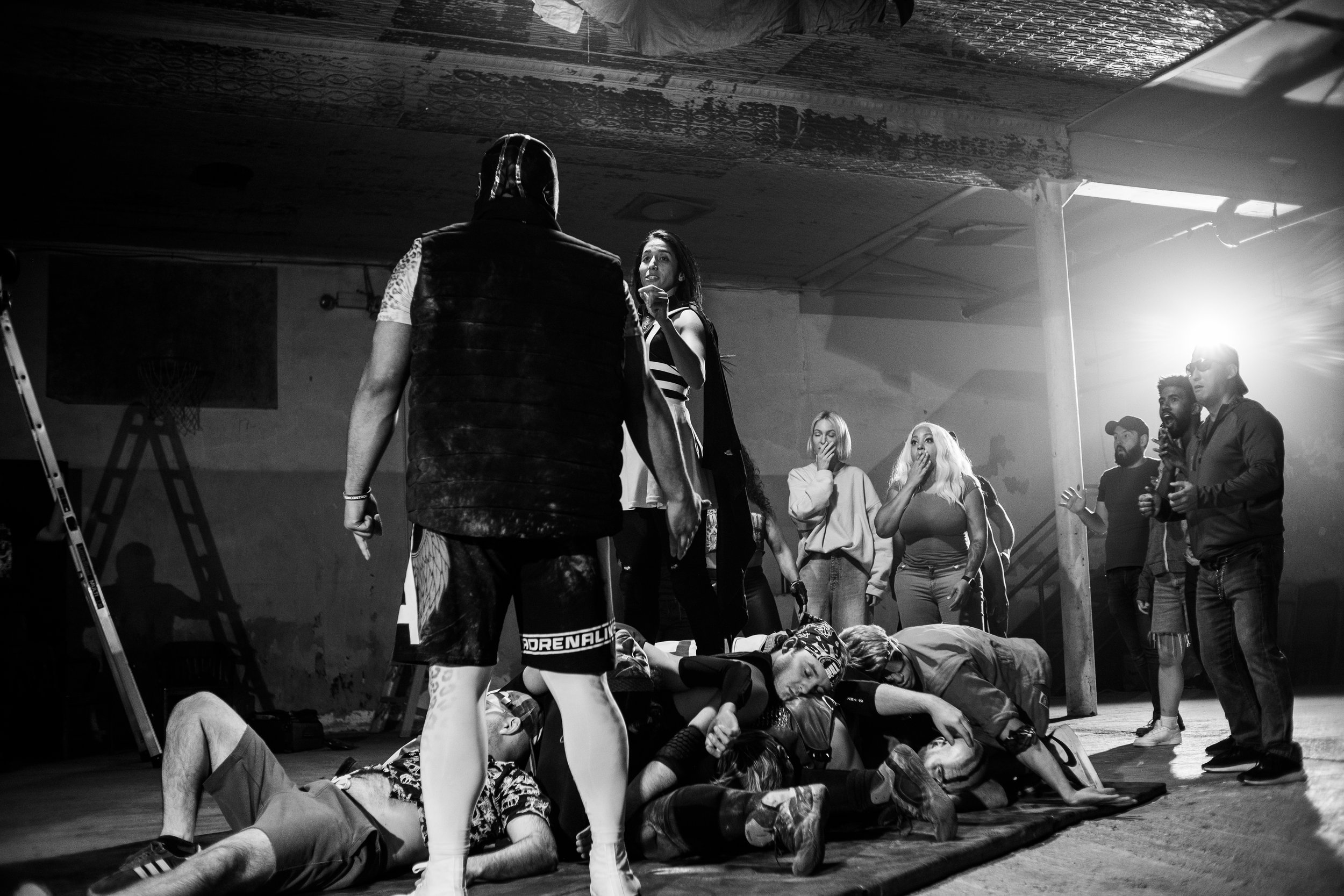 A black and white scene depicting a dramatic moment with people in a room. In the foreground, a masked person stands while several people are on the ground. Other individuals stand in the background observing with shocked expressions. The setting app