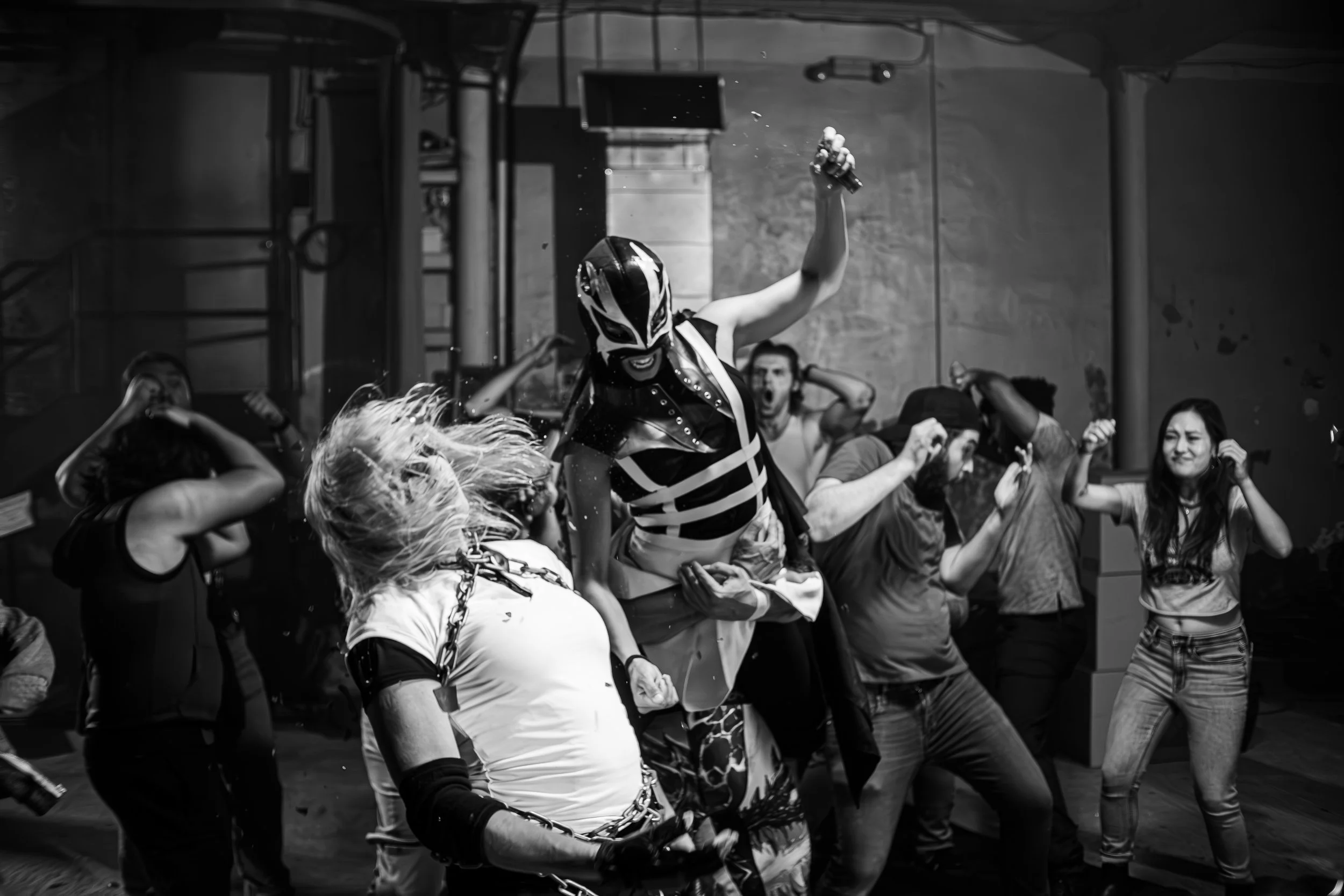 Black and white photo of a lively party scene with people dancing energetically. A central figure wears a costume with a mask, resembling a wrestler. The background shows more people engaged in the event with dynamic expressions.