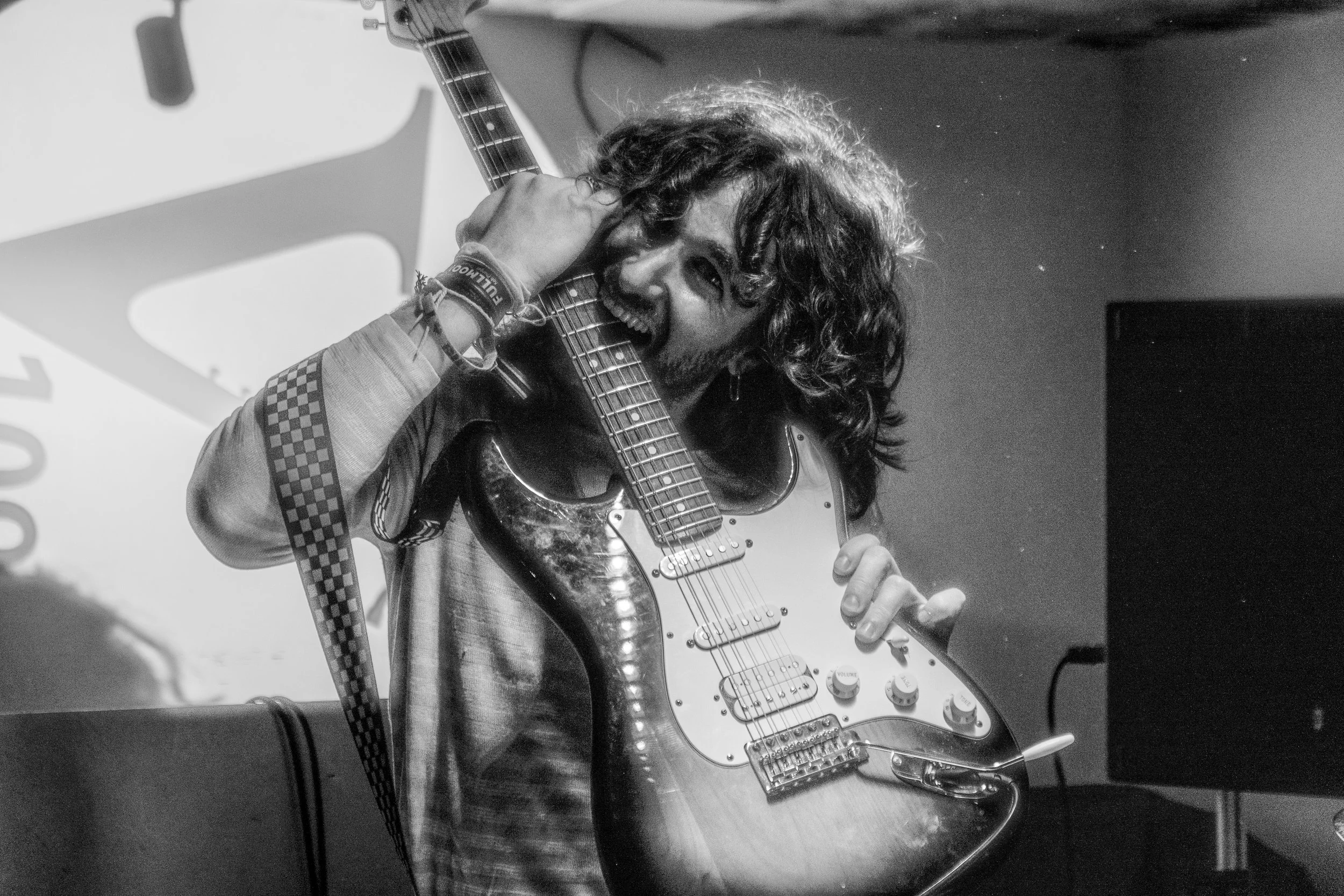 Black and white photo of a person with long hair holding an electric guitar and playfully biting the neck, with a playful and energetic expression.