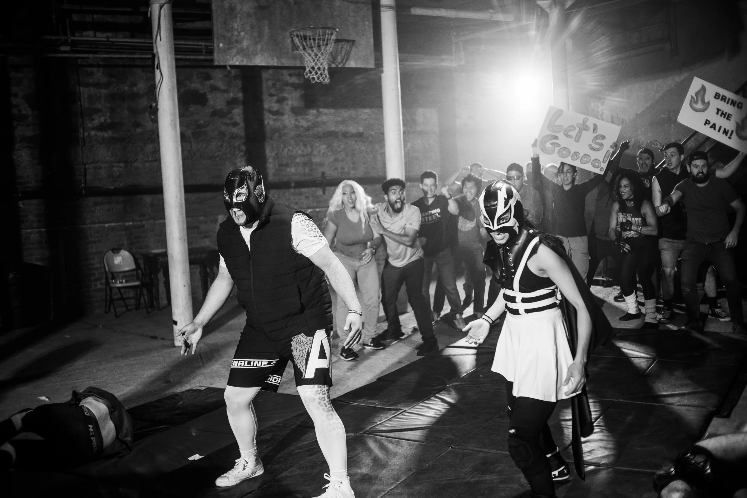 Wrestlers in masks performing for an enthusiastic crowd in a dimly lit indoor space with spectators cheering and holding signs, "Let’s Go!" and "Bring the Pain!"