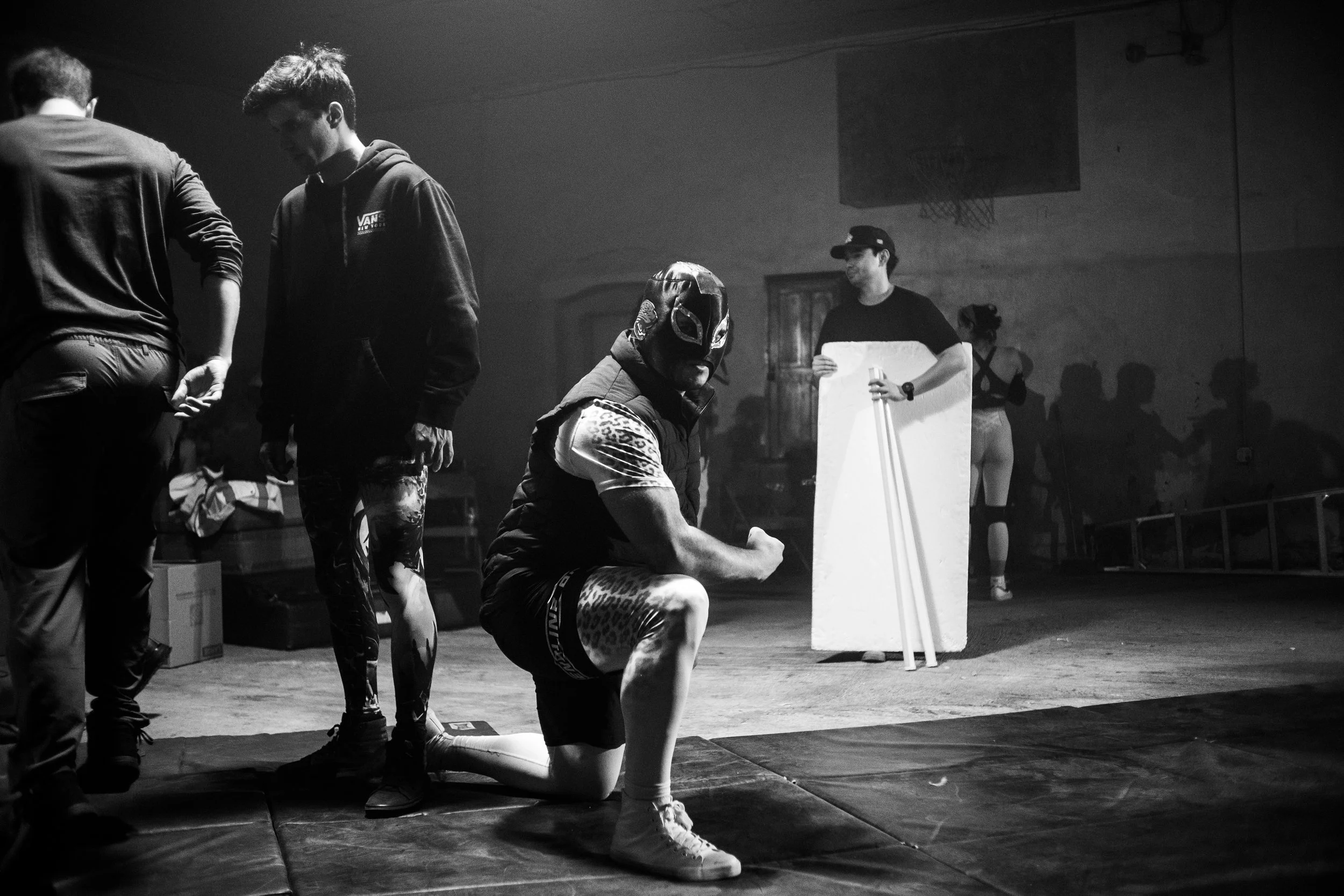 Black and white indoor scene with a man in a wrestling mask kneeling, flexing arm. Others stand around holding equipment, box in background.