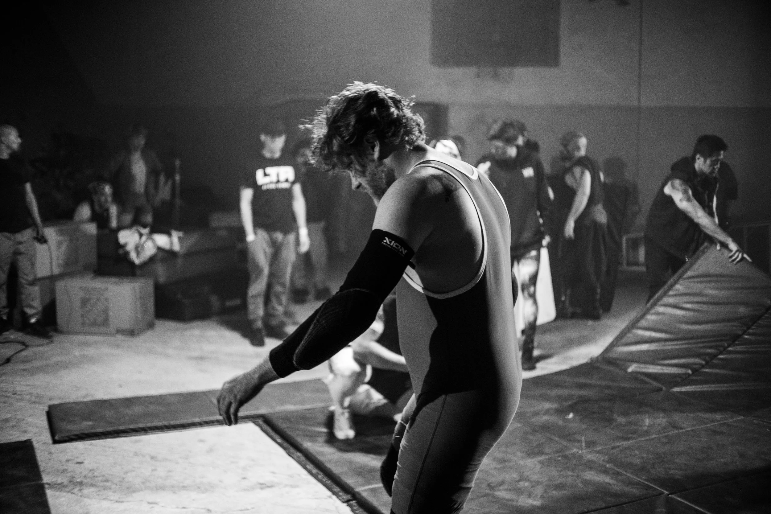Black and white photo of a wrestling event with a focus on a wrestler stretching, surrounded by people and equipment in the background.