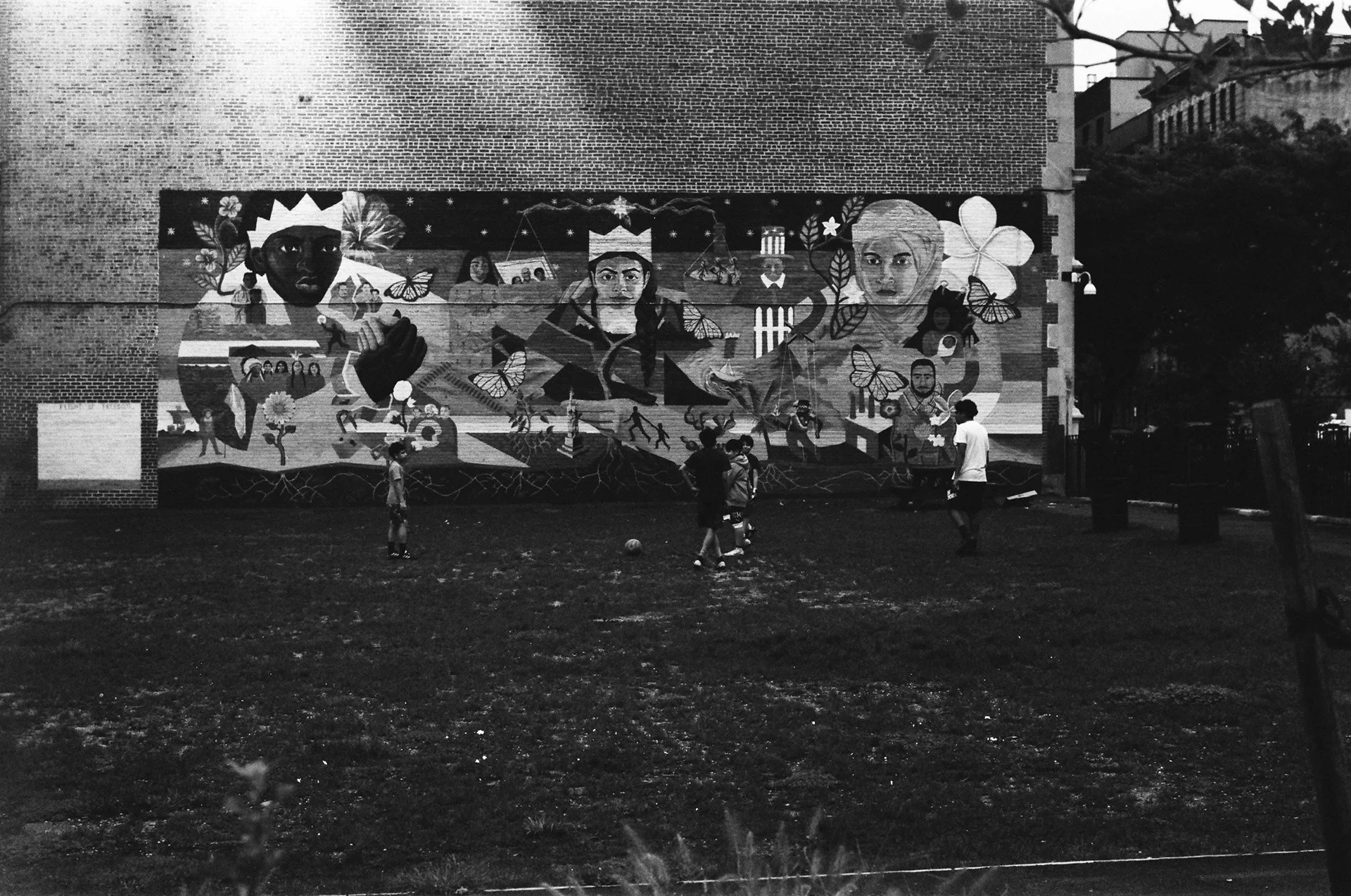 Black and white photo of a brick wall mural with colorful abstract figures and patterns. Several children are playing soccer on the grassy field in front of the mural.
