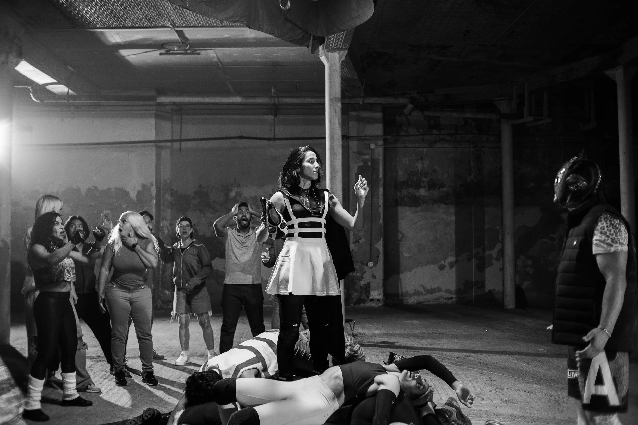 Black and white photo of a woman standing on a floor covered with people, surrounded by a group of spectators. The scene is dramatic, likely in an industrial setting.