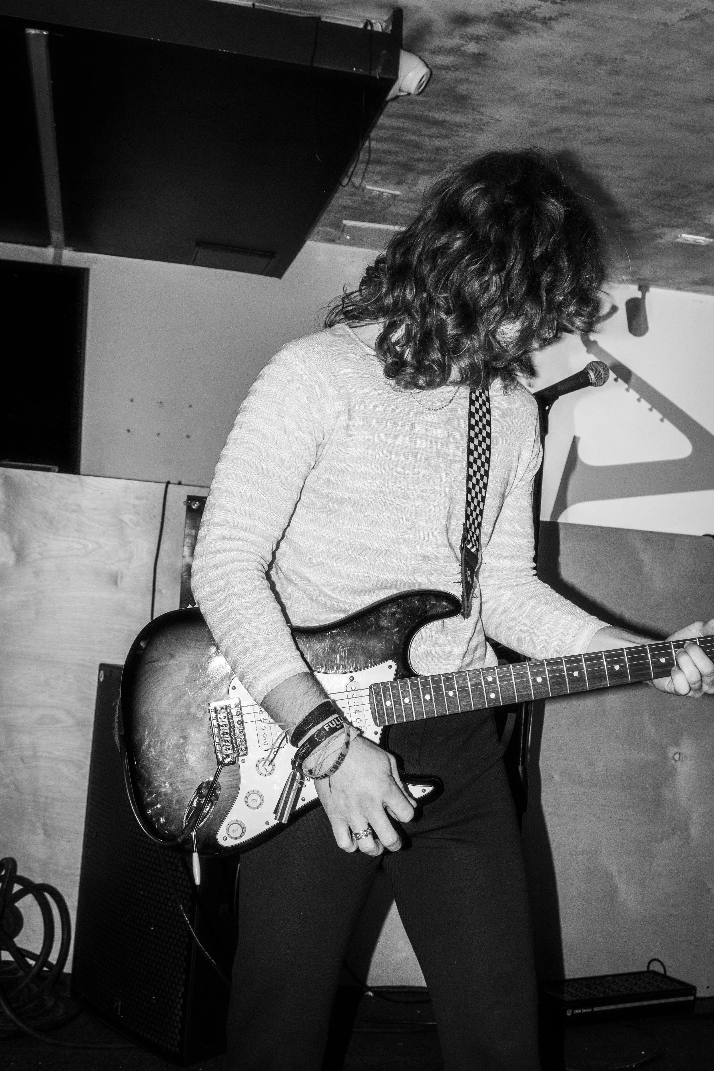 Black and white photo of a person with long hair playing an electric guitar, standing near a microphone in a room with a low ceiling.