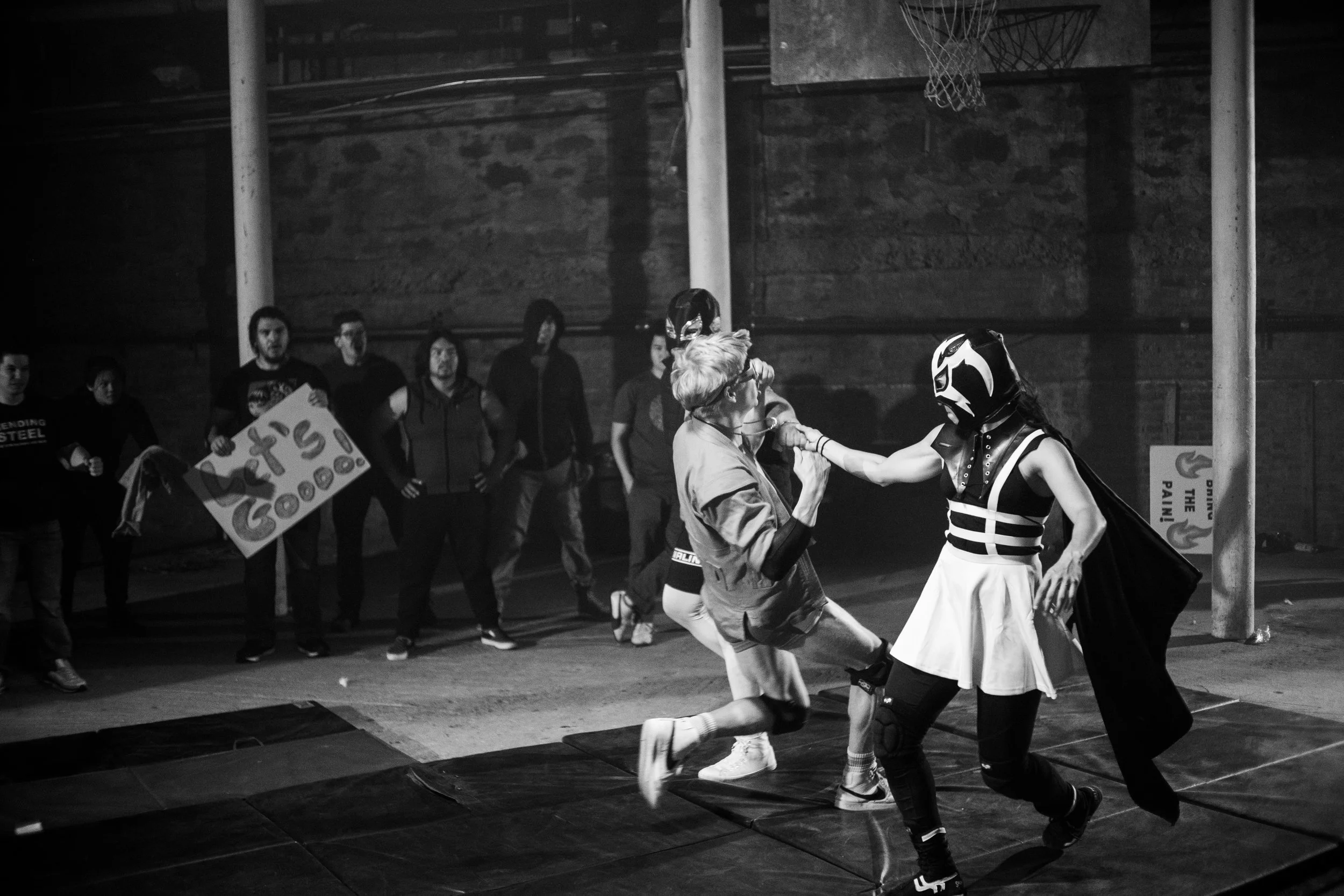 Masked wrestler performing a move on an opponent in an underground wrestling match, with spectators watching and holding a sign saying "Let's Go!"
