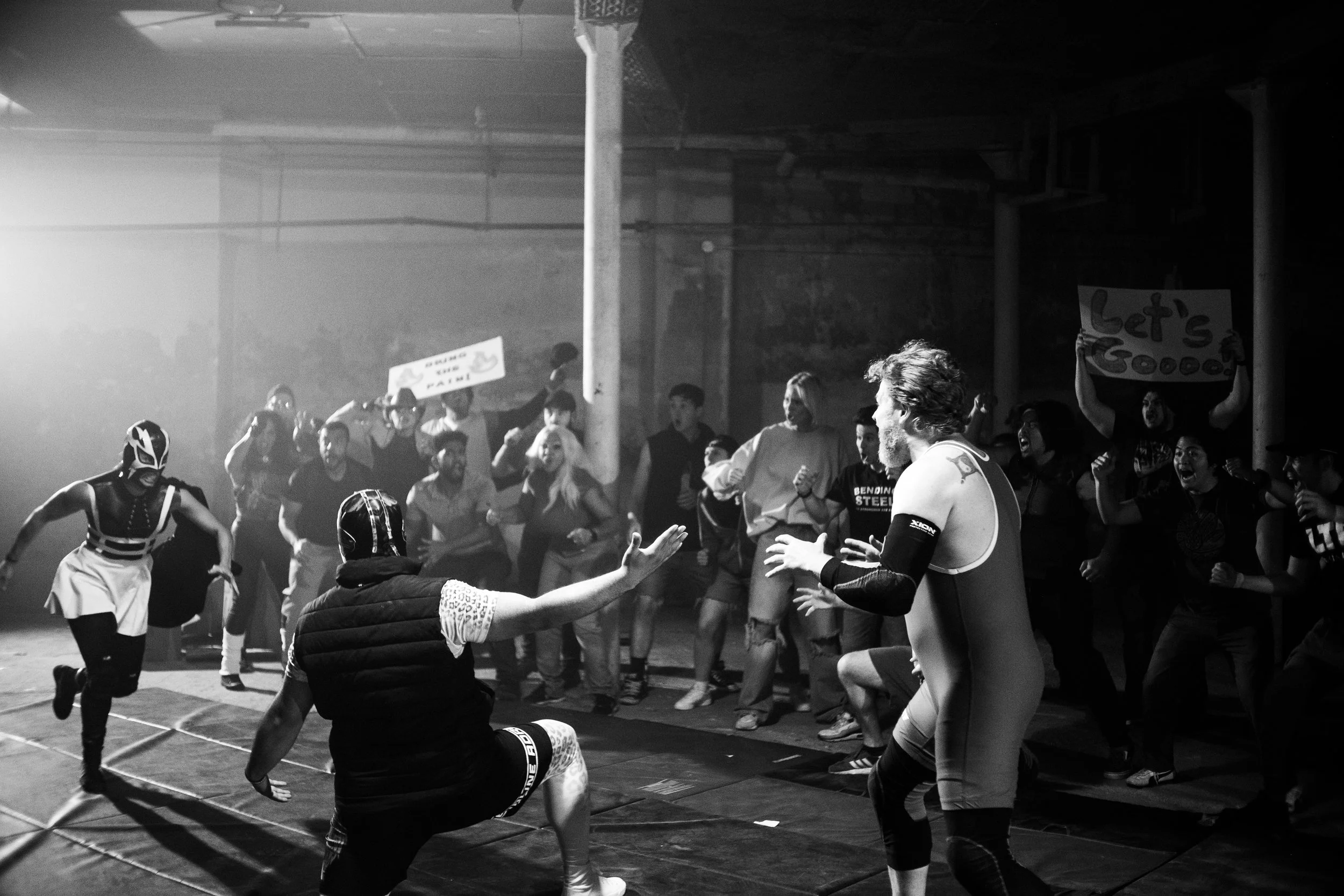 Black and white image of a wrestling match with masked wrestlers in an arena, surrounded by a cheering crowd holding signs.