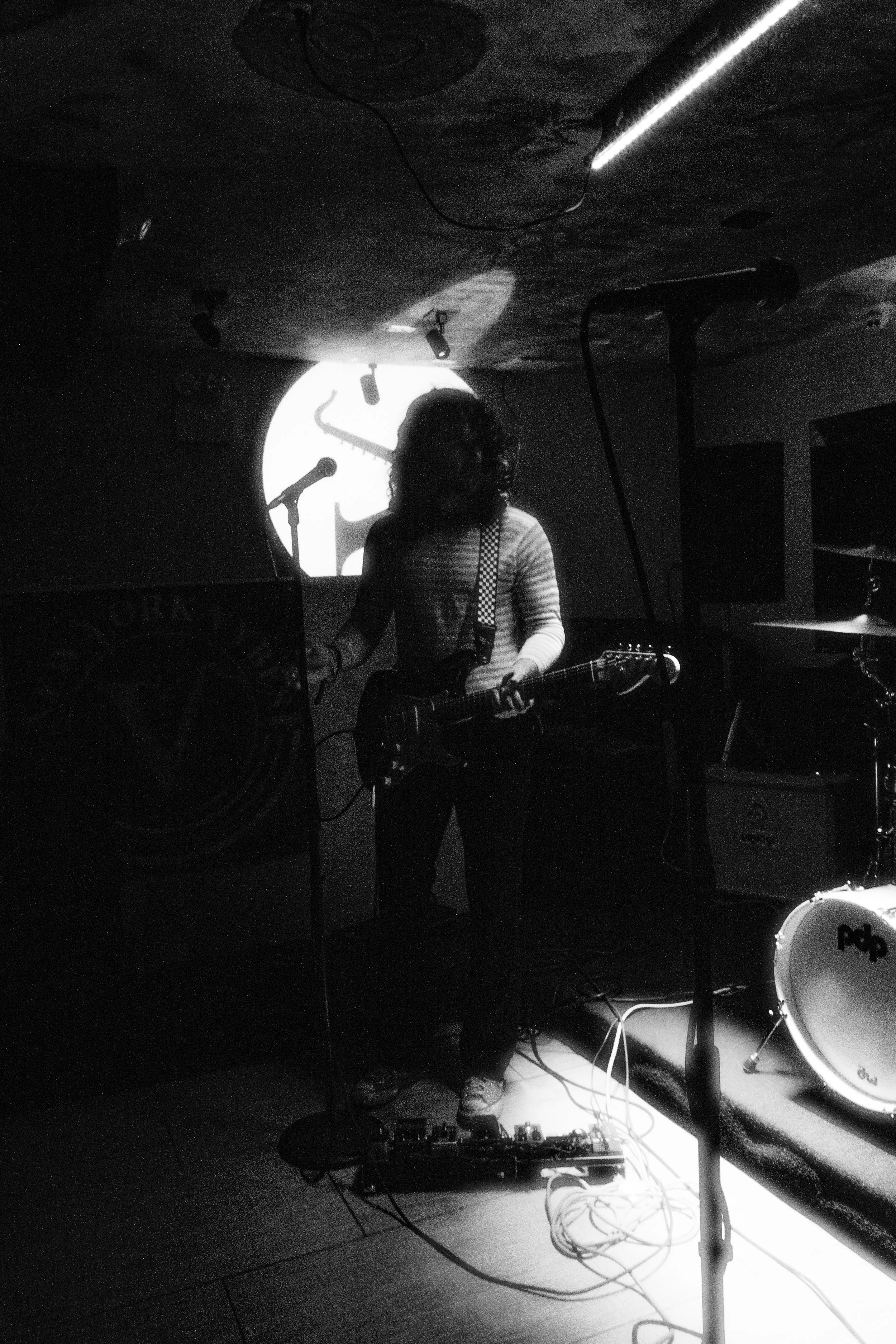 Black and white image of a musician playing an electric guitar on stage, with a microphone and drum kit nearby. A spotlight casts a shadow on the backdrop.