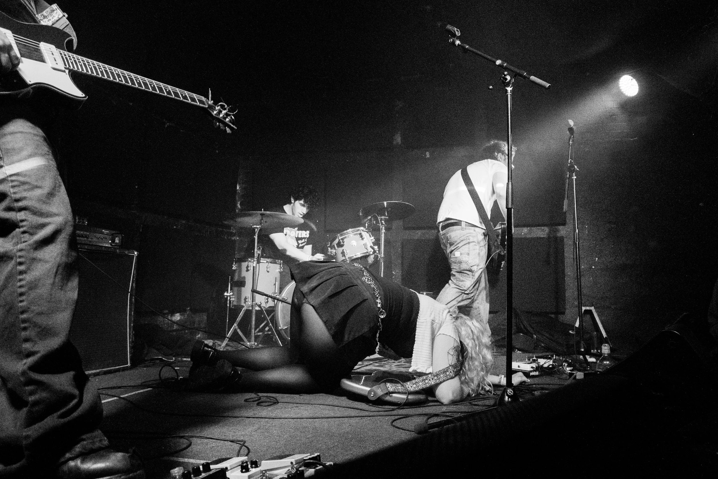 Black and white photo of a live band performance with musicians playing guitar and drums on stage, one female musician lying on the ground.