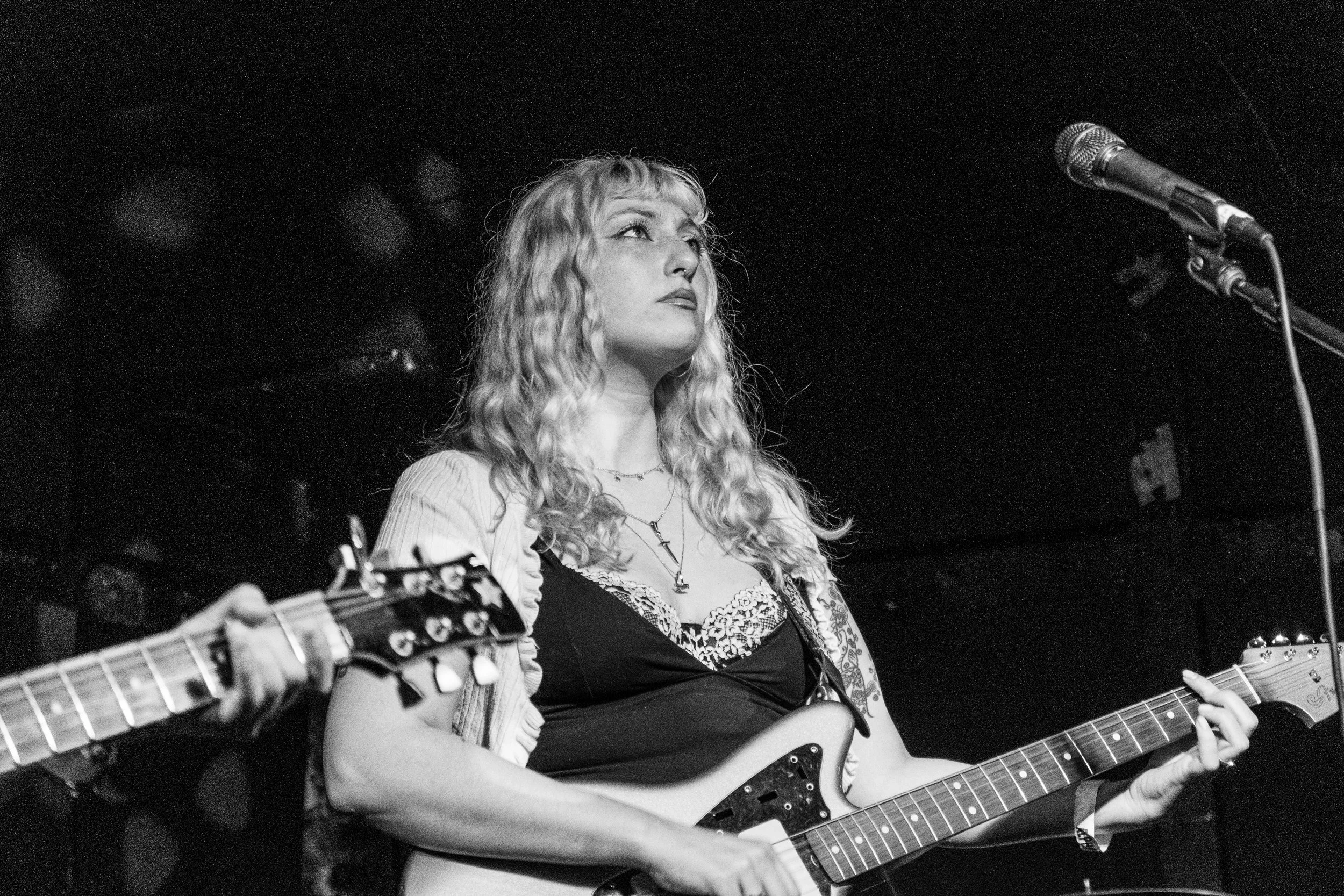 Black and white photo of a musician with long hair playing an electric guitar on stage beside a microphone.