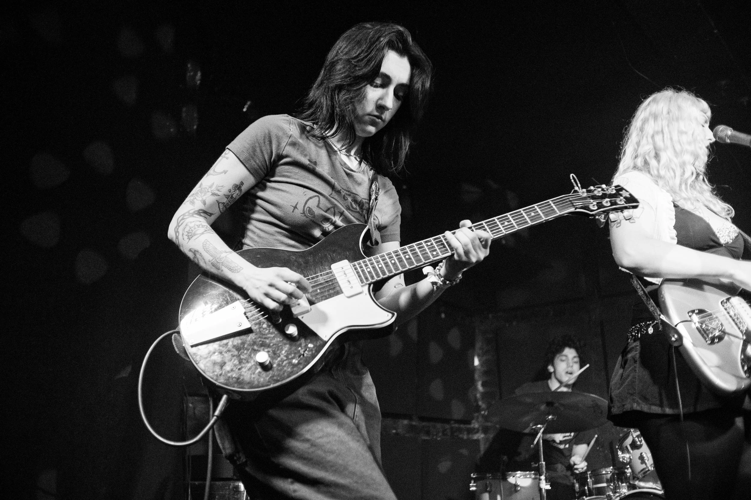Black and white photo of a musician playing an electric guitar on stage, with a drummer and another musician in the background.