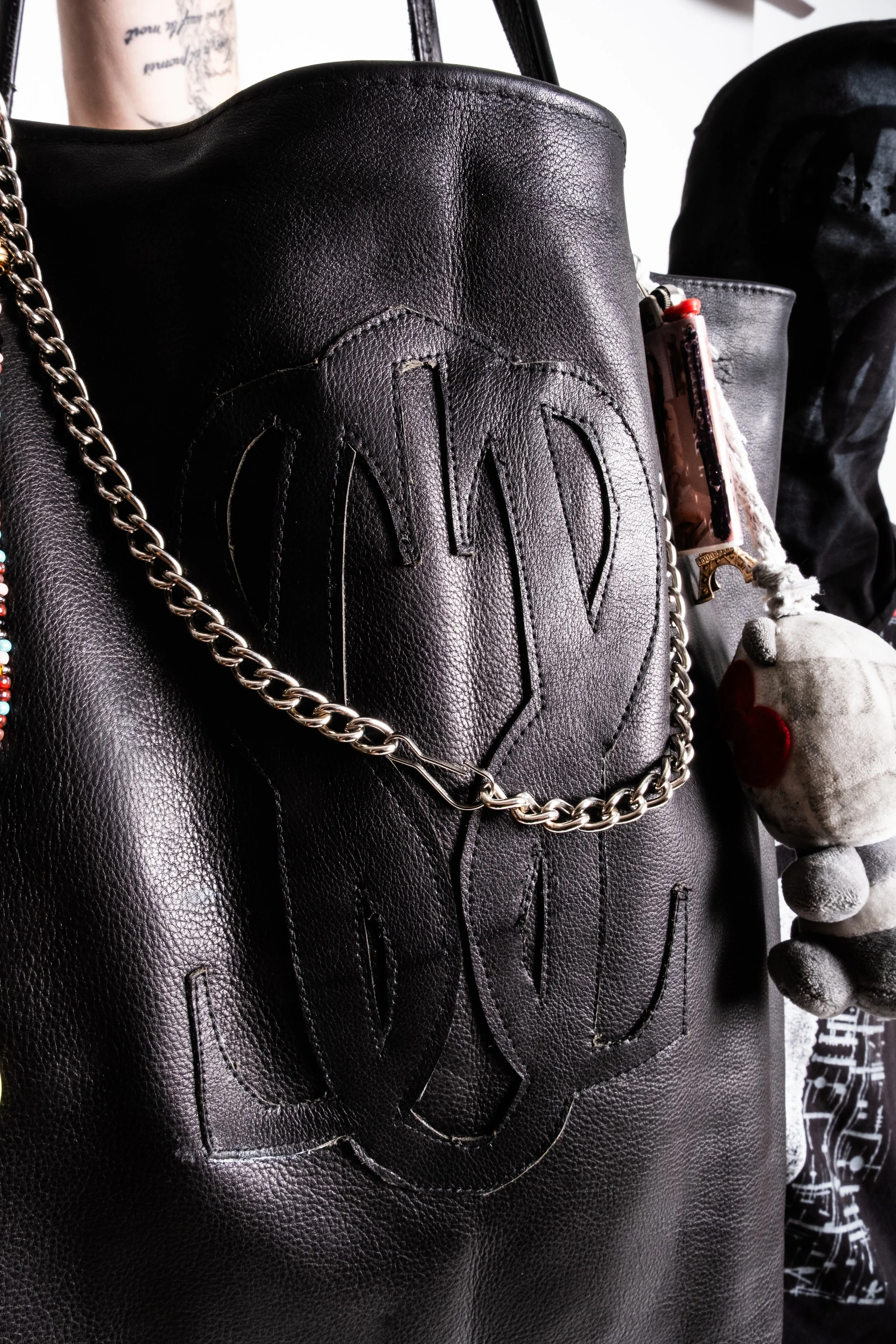 Close-up of a black leather handbag with decorative stitching and a metal chain, featuring a plush toy keychain hanging from the side.