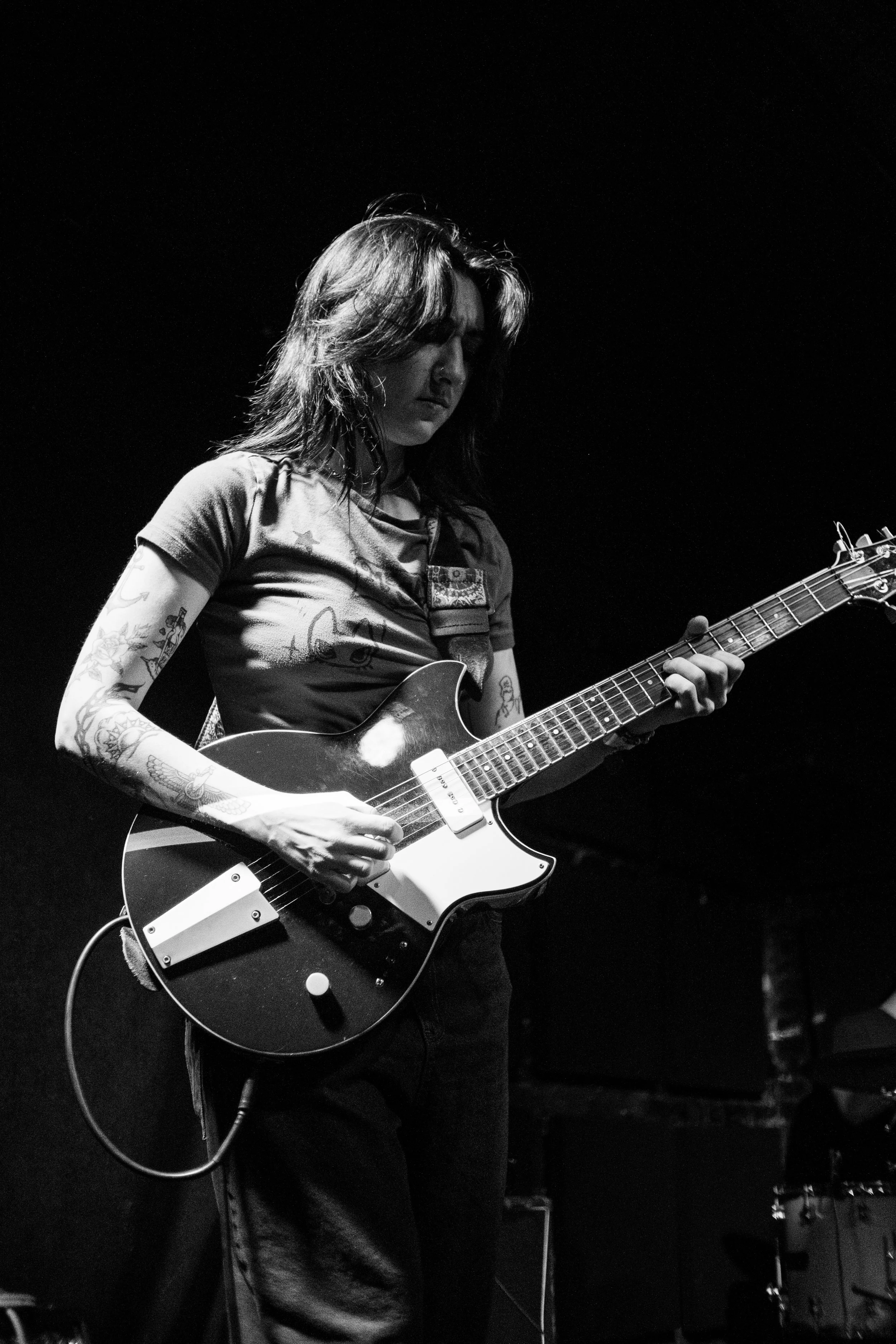 Black and white photo of a person with long hair and tattoos playing an electric guitar on stage.