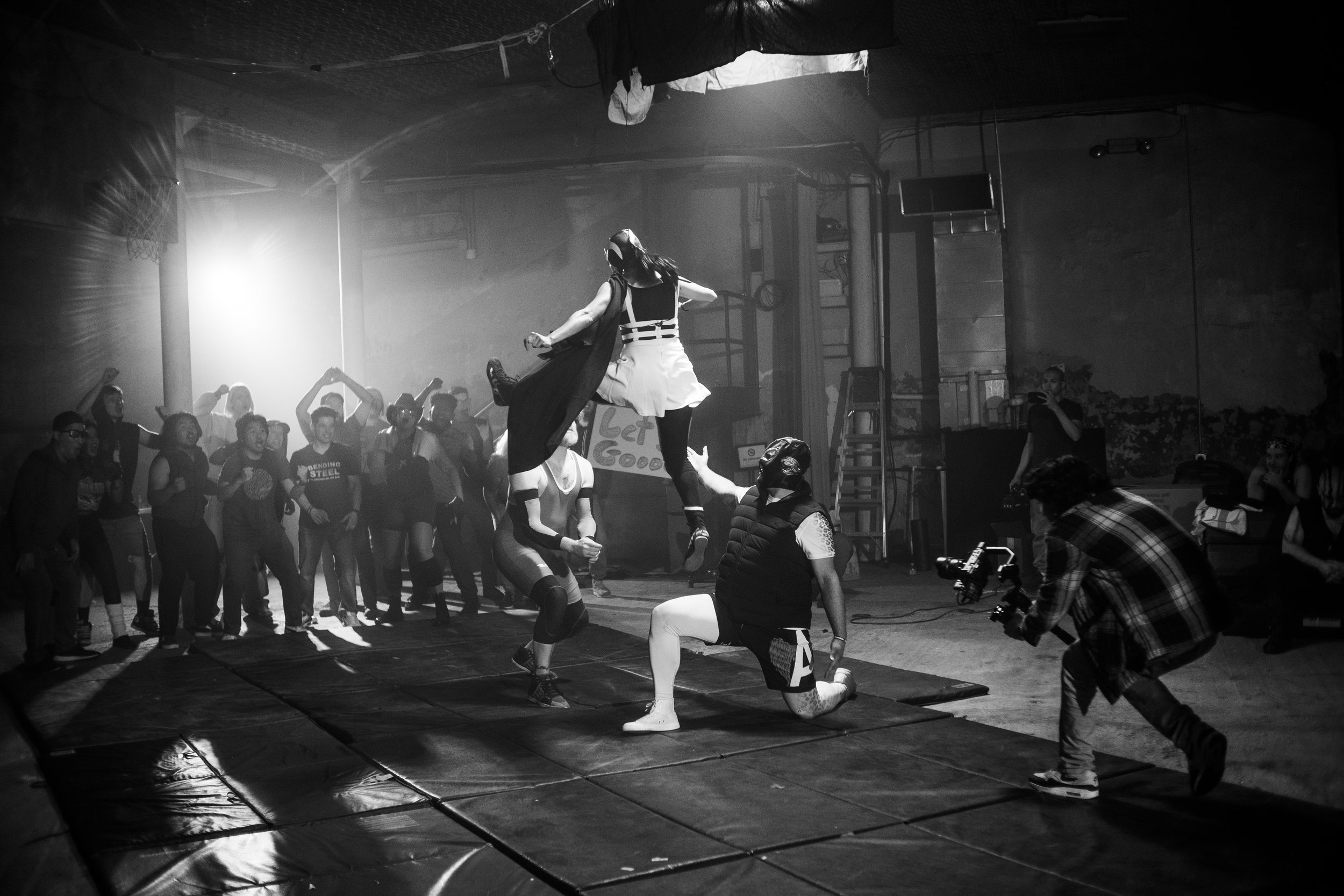 Black and white photo of an indoor wrestling event with a crowd watching. A wrestler in mid-air performing a move onto another wrestler. A camera operator is capturing the action.