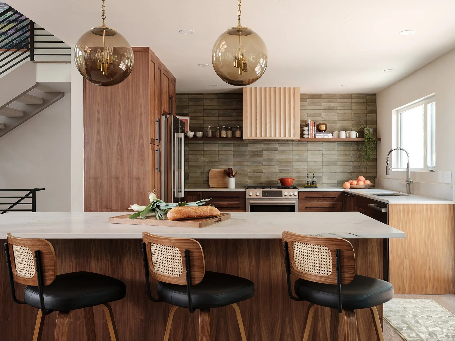 Modern kitchen with wooden cabinets, marble island, and bar stools, featuring pendant lights, a stove, and a window over the sink.