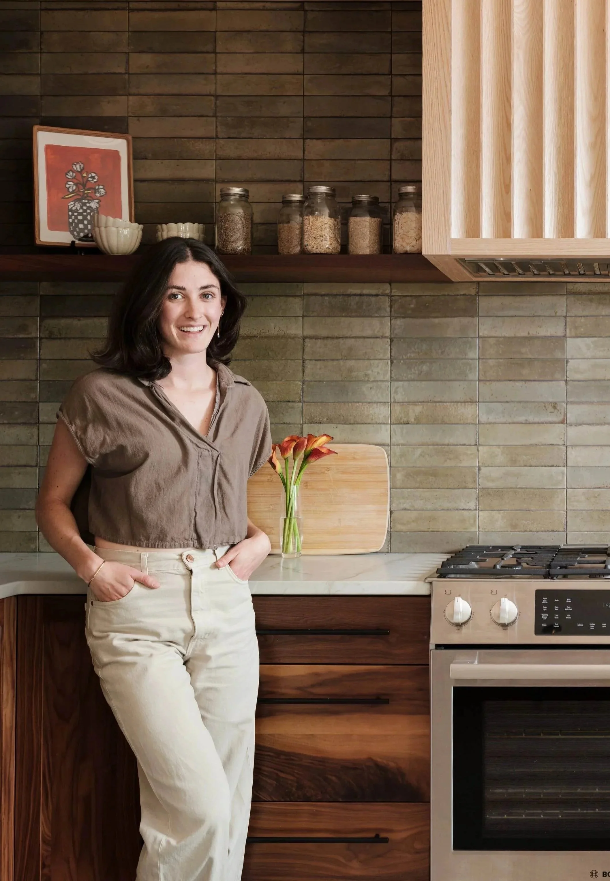 A woman with dark hair smiling, wearing a beige crop top and cream-colored pants, standing in a modern kitchen with brown wooden cabinets, gray tile backsplash, glass jars of spices, a framed artwork, and a vase with orange flowers on the countertop.