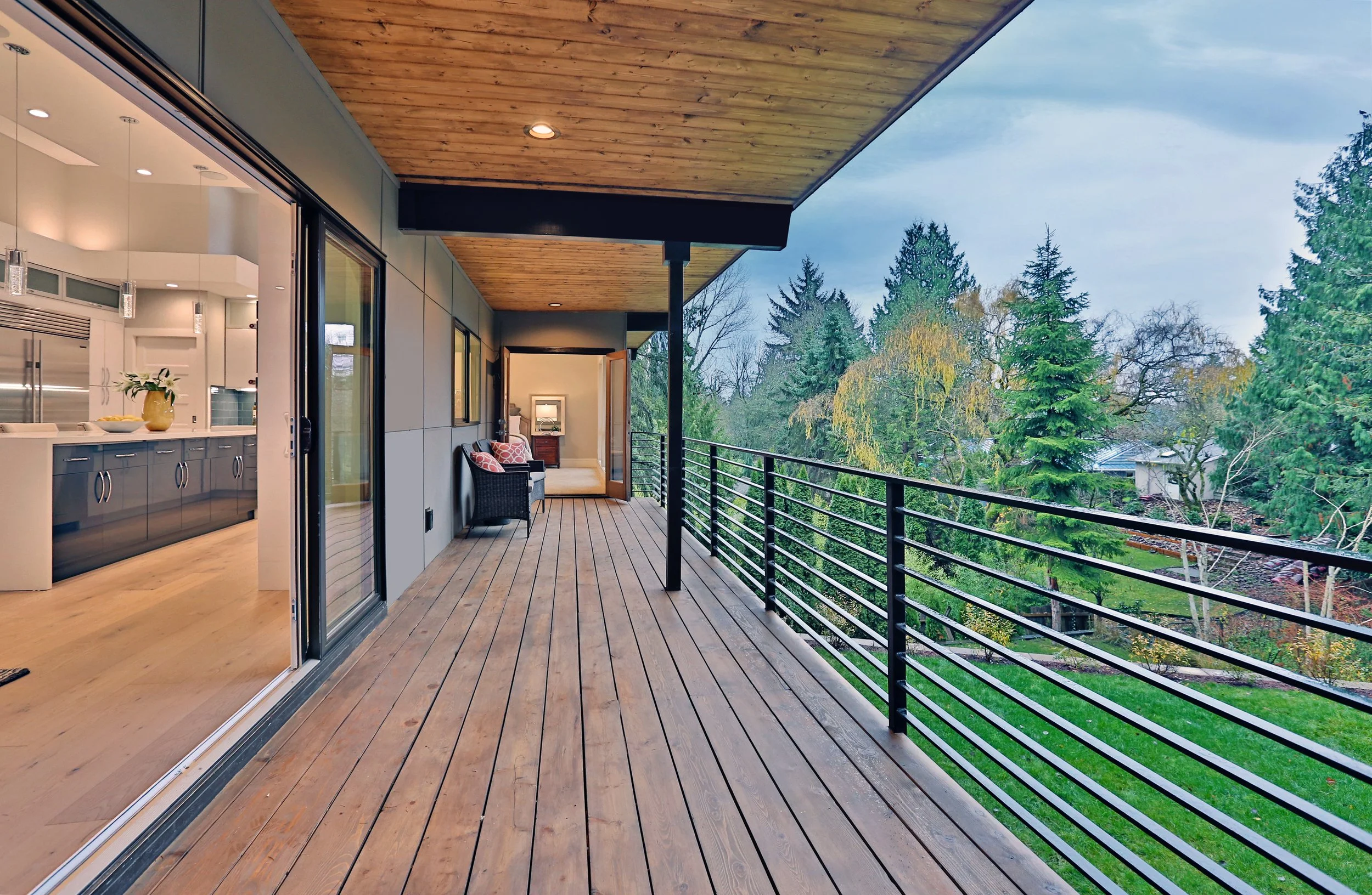 Balcony with wooden flooring and ceiling, partial indoor view of kitchen with blue cabinets, and lush green trees and houses in the background.