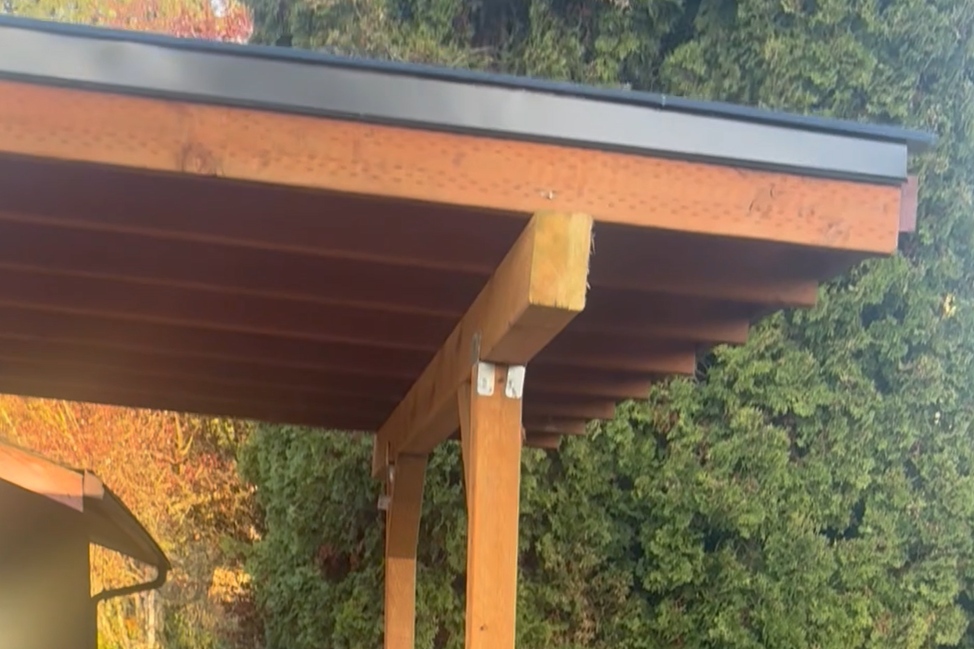Close-up of a wooden deck or porch ceiling with an overhanging roof, supported by a wooden post, surrounded by trees with autumn foliage.
