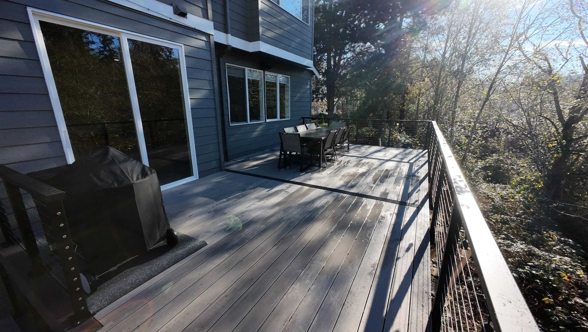 An outdoor wooden balcony with a dining table and six chairs, a barbecue grill, and a view of trees with sunlight shining through.