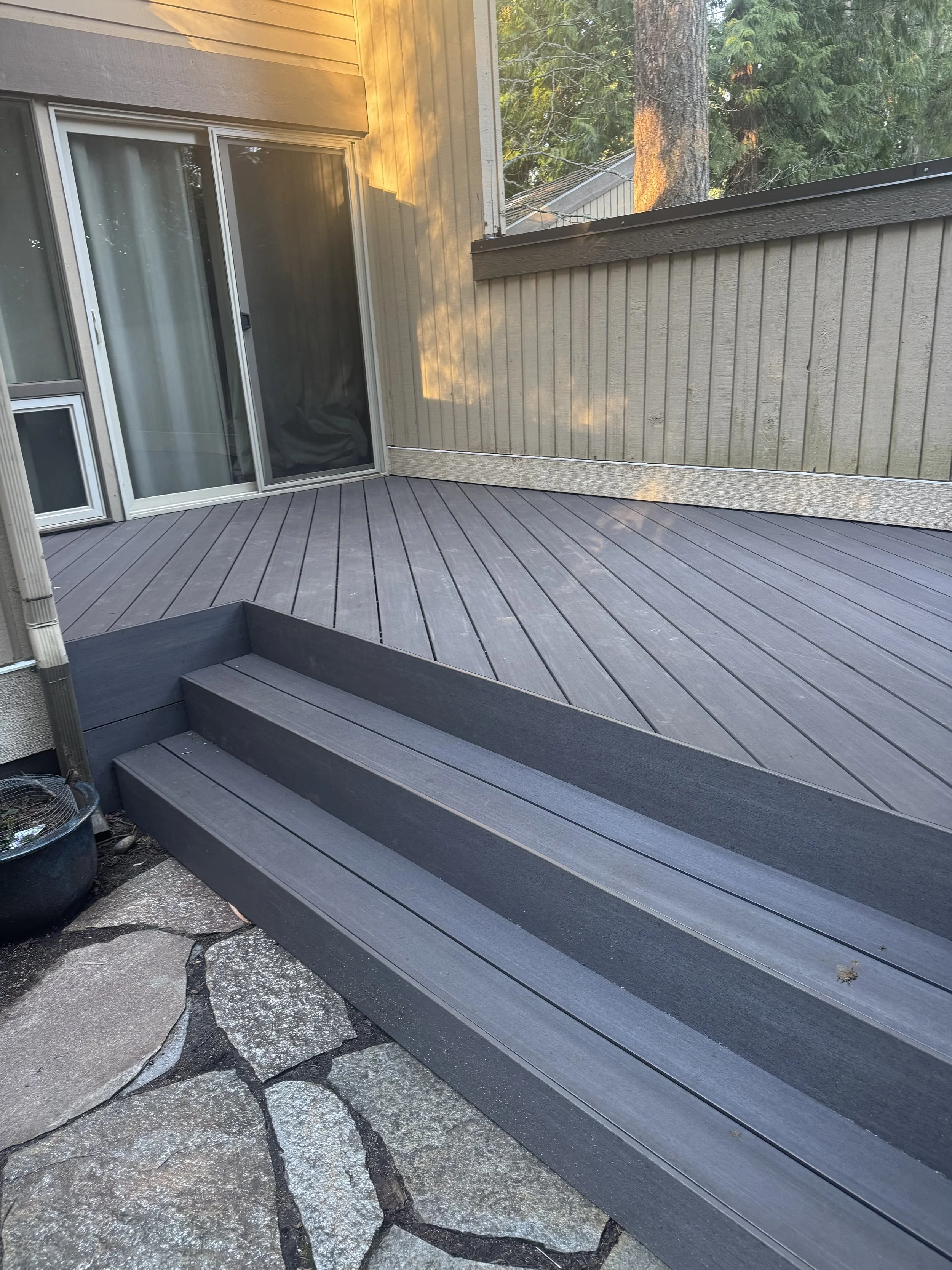 A backyard deck with gray composite wood, leading to sliding glass doors and a vinyl fenced yard. A large tree and greenery are visible in the background.