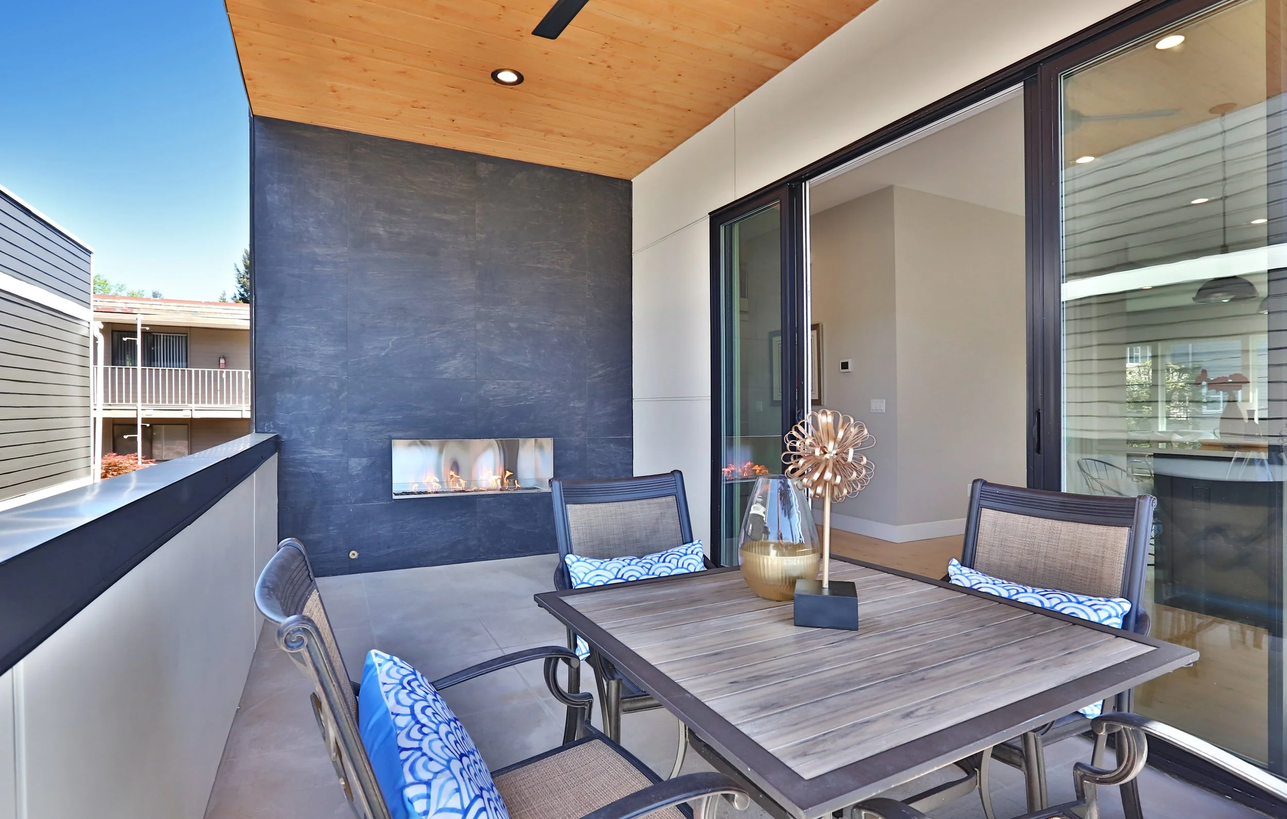 Balcony with a wooden ceiling, black tile wall, and a modern gas fireplace. There is a dining table with chairs, decorative pillows, a gold vase, and sliding glass doors leading inside.