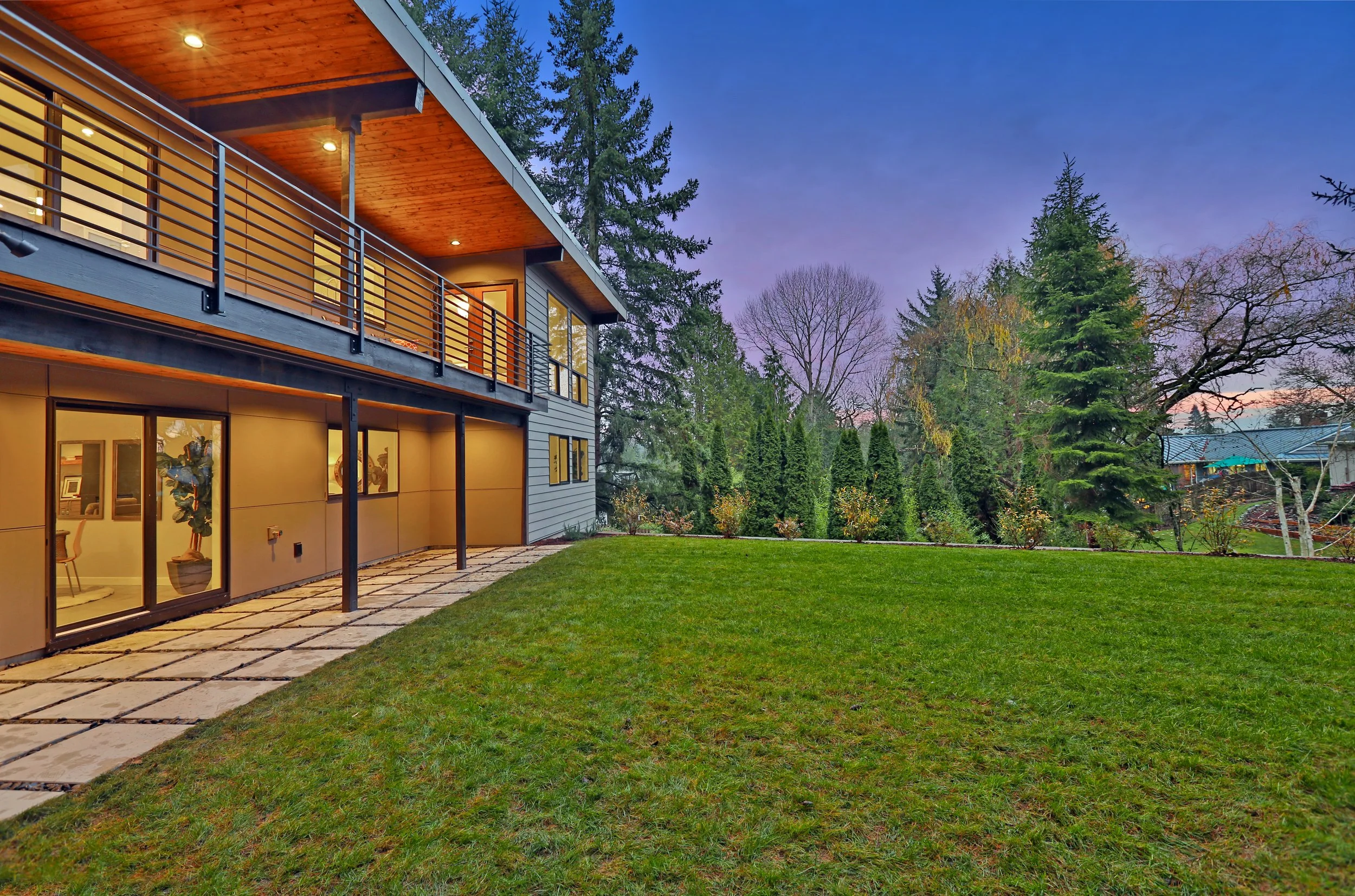 Modern two-story house with a large backyard, green grass, trees, and a wooden ceiling over a balcony at dusk.