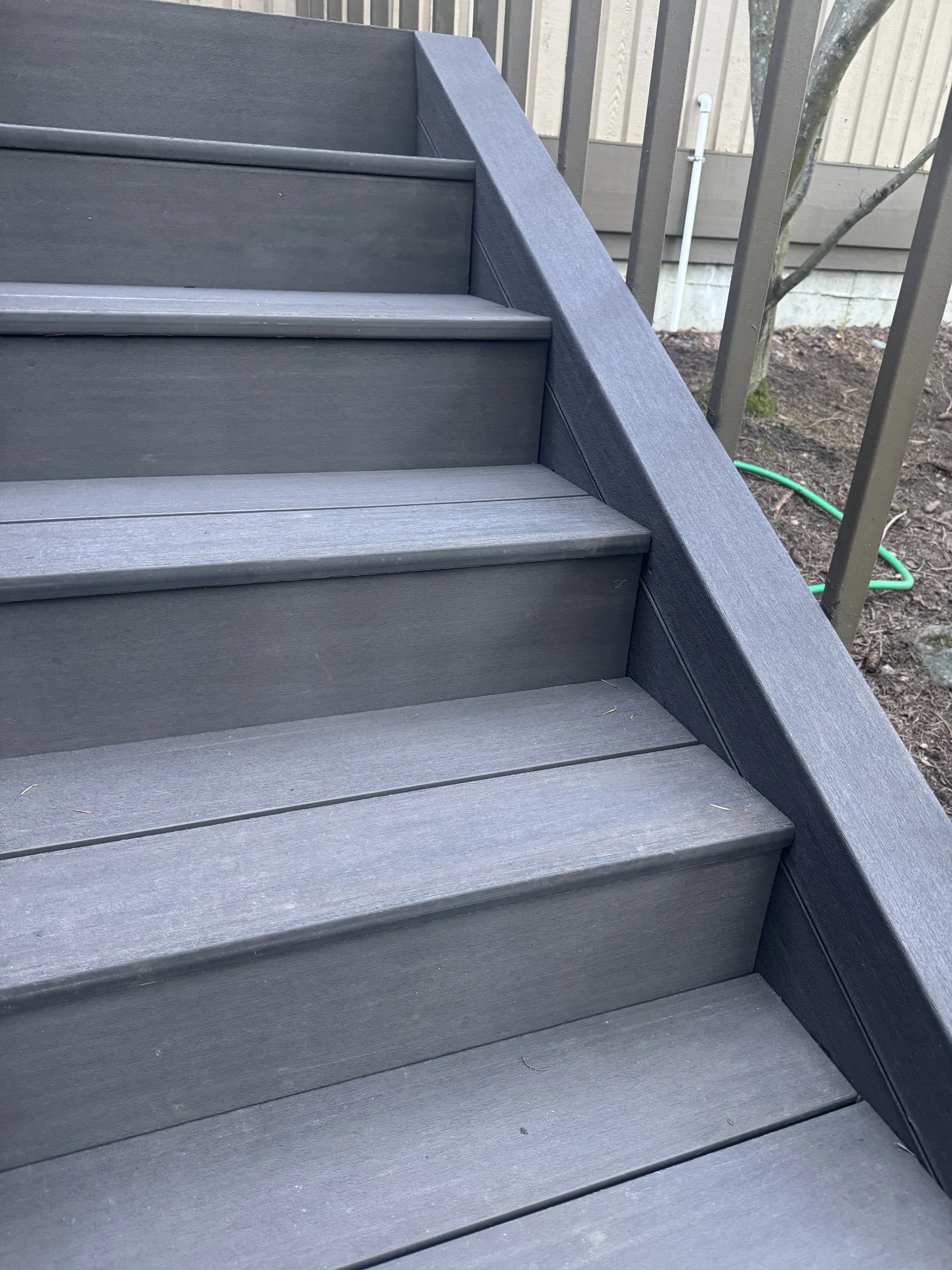 Close-up view of outdoor gray wooden stairs with a slope and metal railing, adjacent to a garden area with soil and a tree, and a green garden hose on the ground.