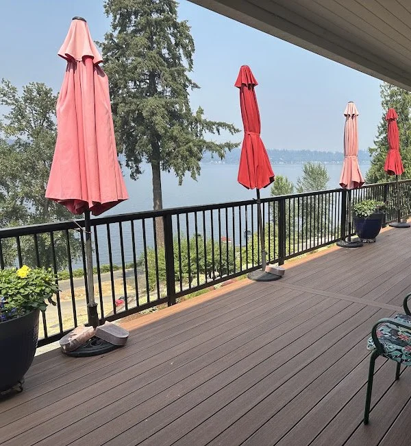 A balcony with four closed red umbrellas, potted plants, and a view of a lake with trees in the background.