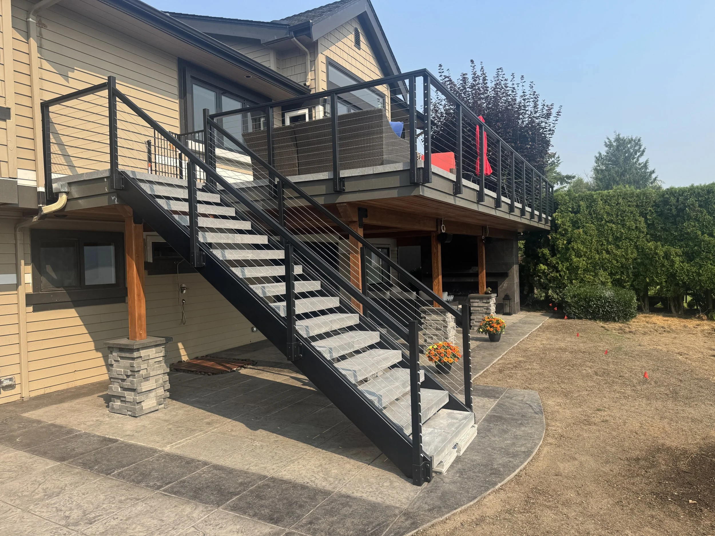 A house with a black metal staircase leading to a balcony with outdoor furniture and red umbrellas, surrounded by trees and a landscaped yard.
