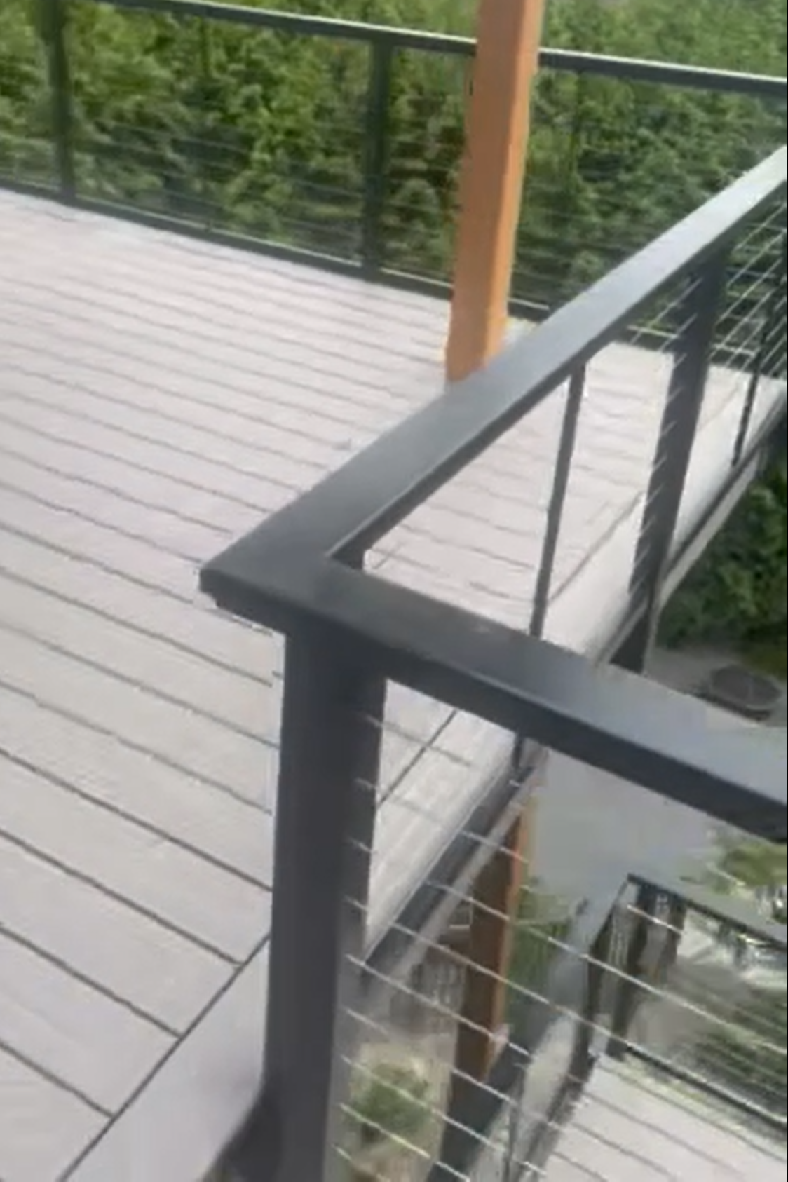 View of a balcony with light-colored wooden flooring, black metal railing with horizontal wires, and a wooden post, overlooking green trees.
